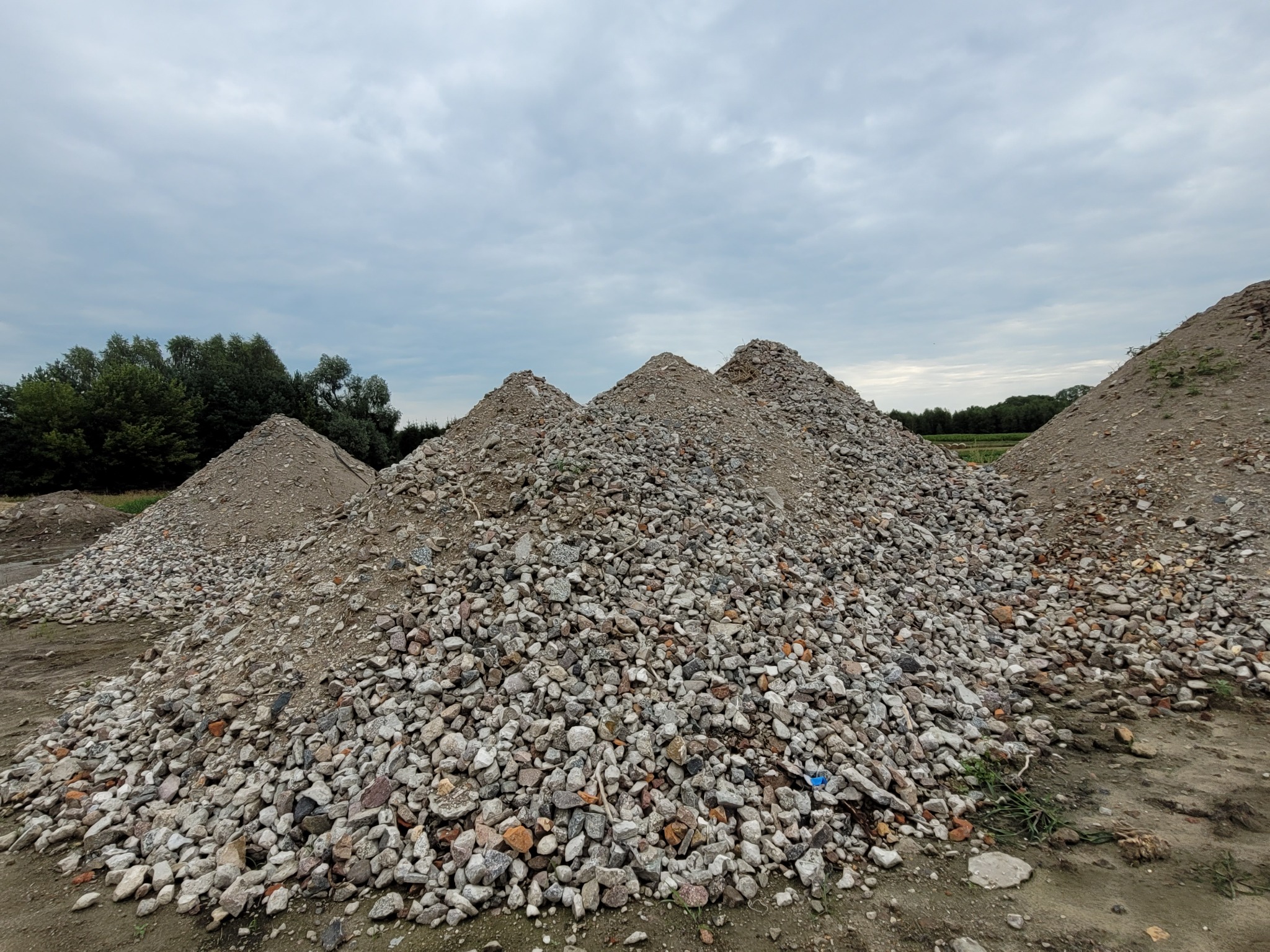 Kupa kamieni i gruzu z fragmentami cegieł ułożona w stożki na placu budowy, z widocznymi drzewami i pochmurnym niebem w tle.