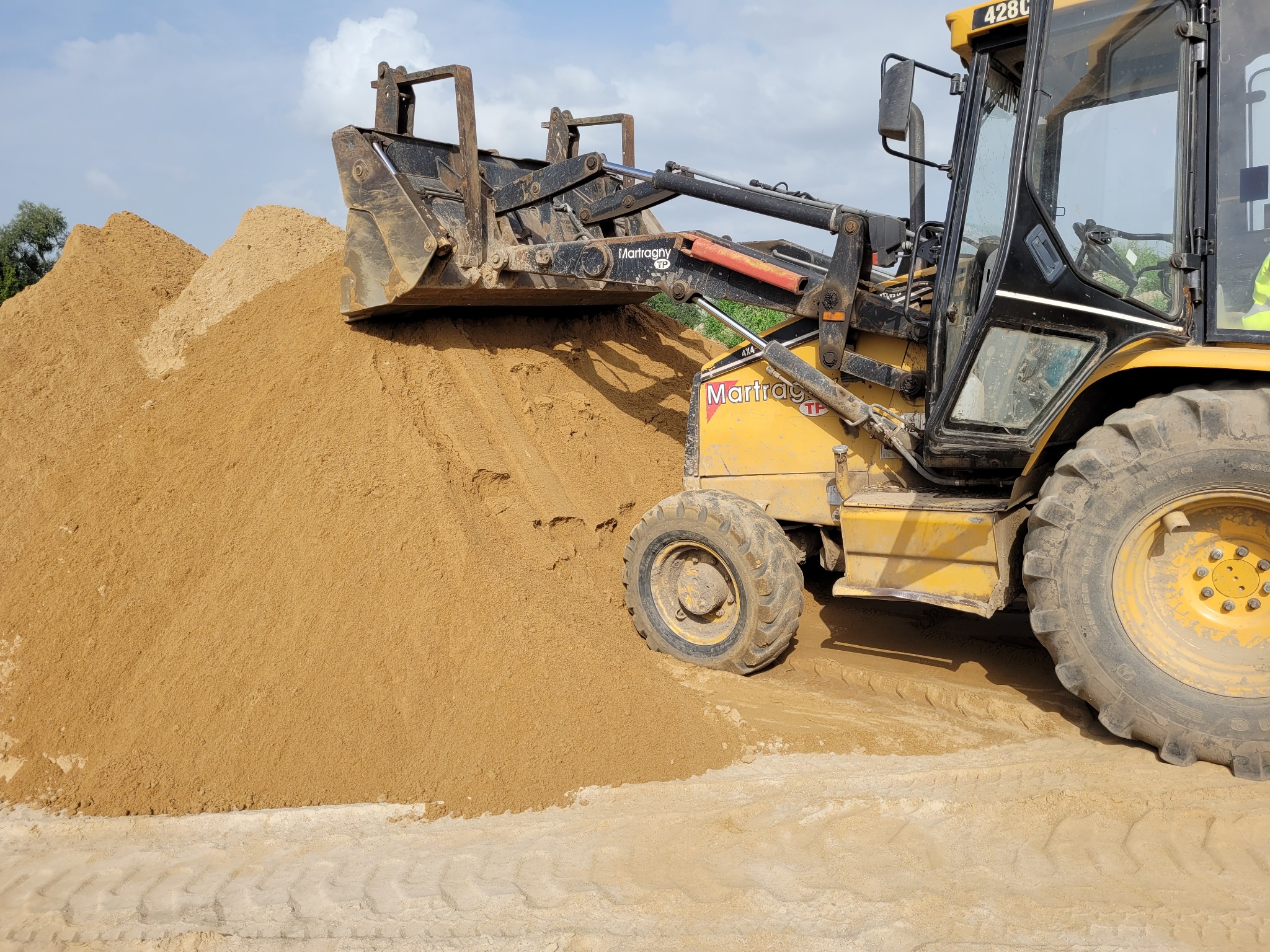Żółta ładowarka czołowa Martragny TP z pełną łyżką piasku na tle dużej pryzmy, widoczne ślady opon na podłożu.