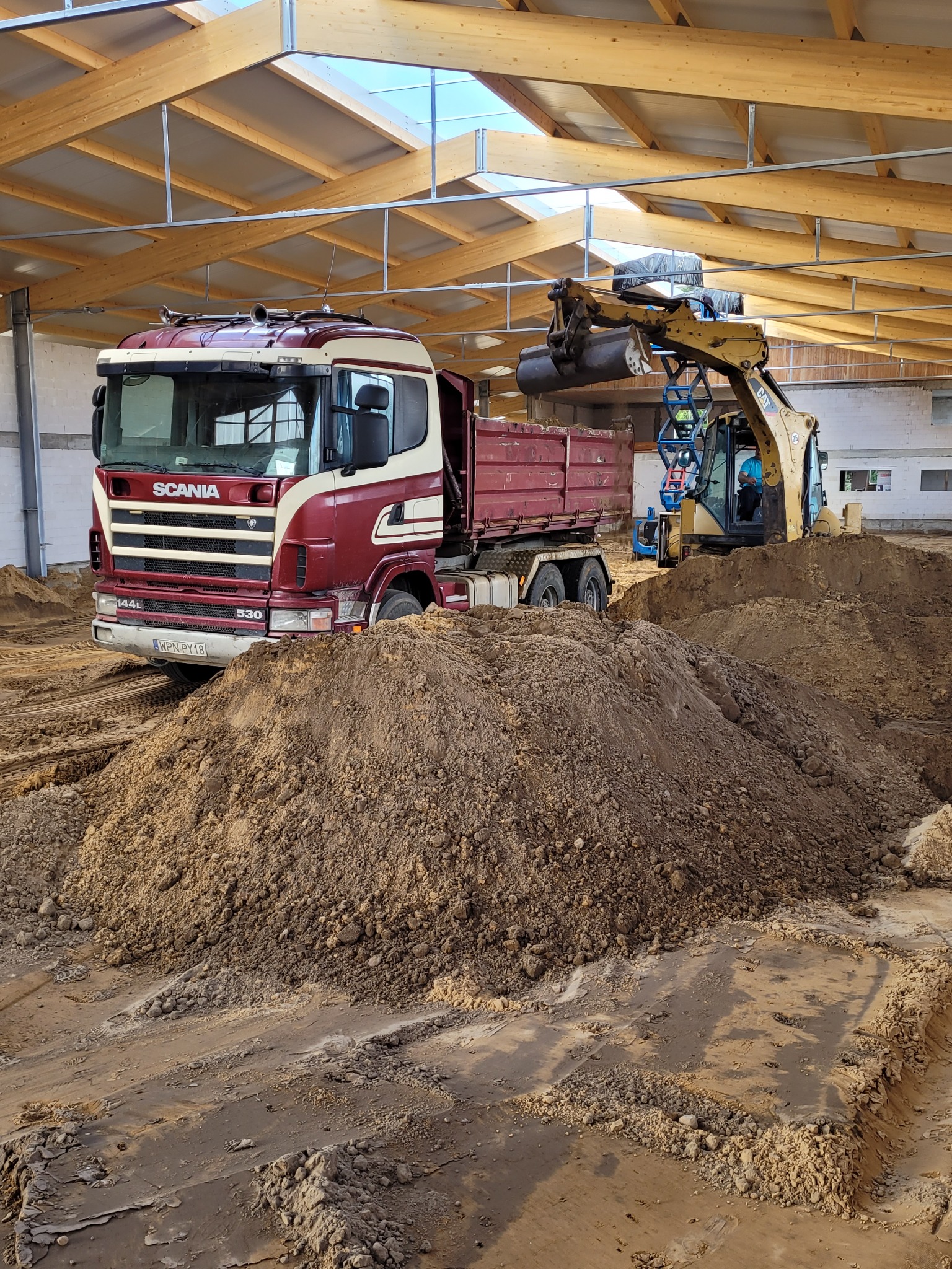 Ciężarówka Scania 144L 530 i koparka CAT 302.5C pracują wewnątrz hali z drewnianą konstrukcją dachu, przesypując ziemię.