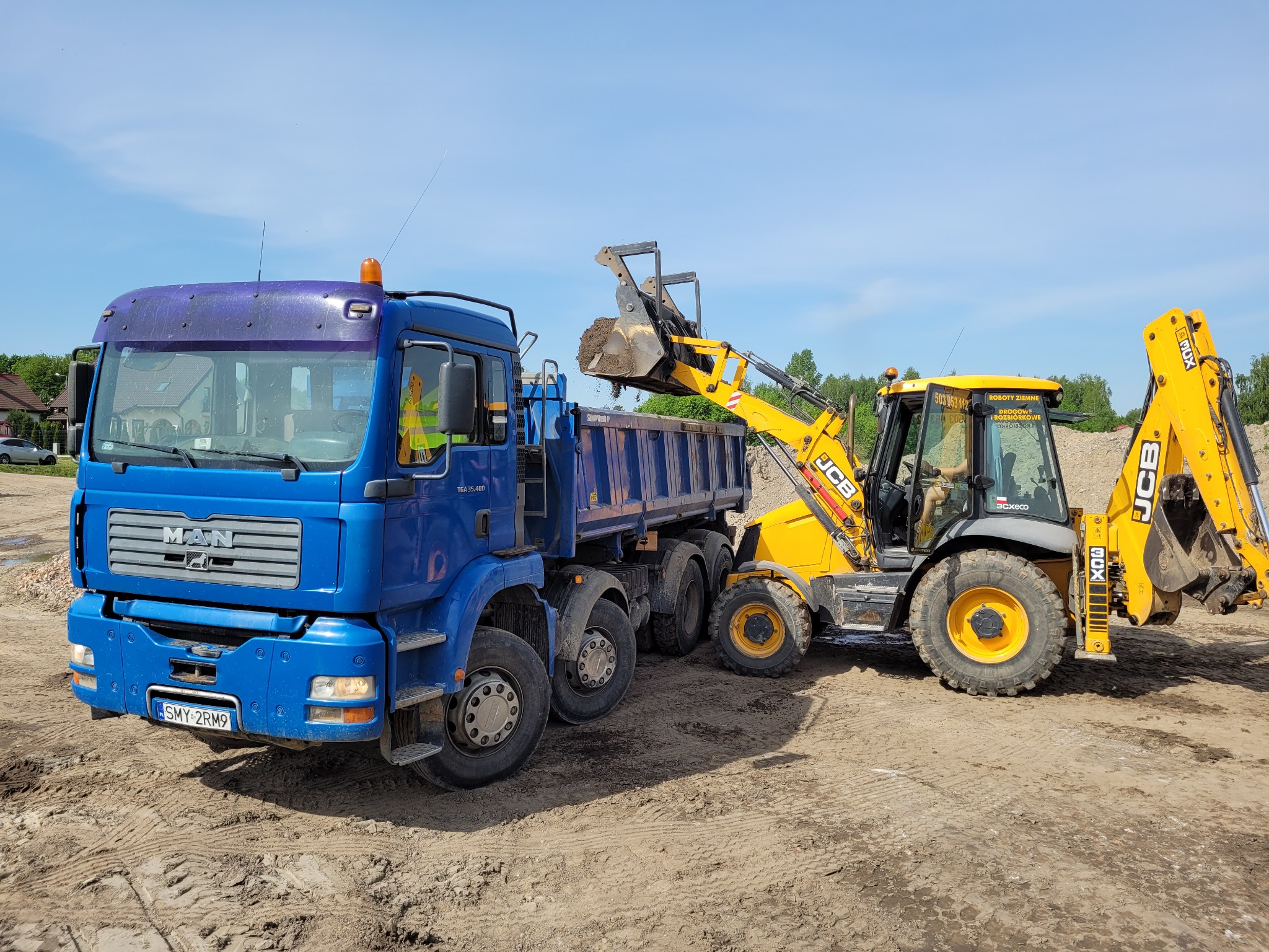 Żółta koparko-ładowarka JCB załadowuje urobek do niebieskiej ciężarówki MAN na placu budowy w słoneczny dzień.