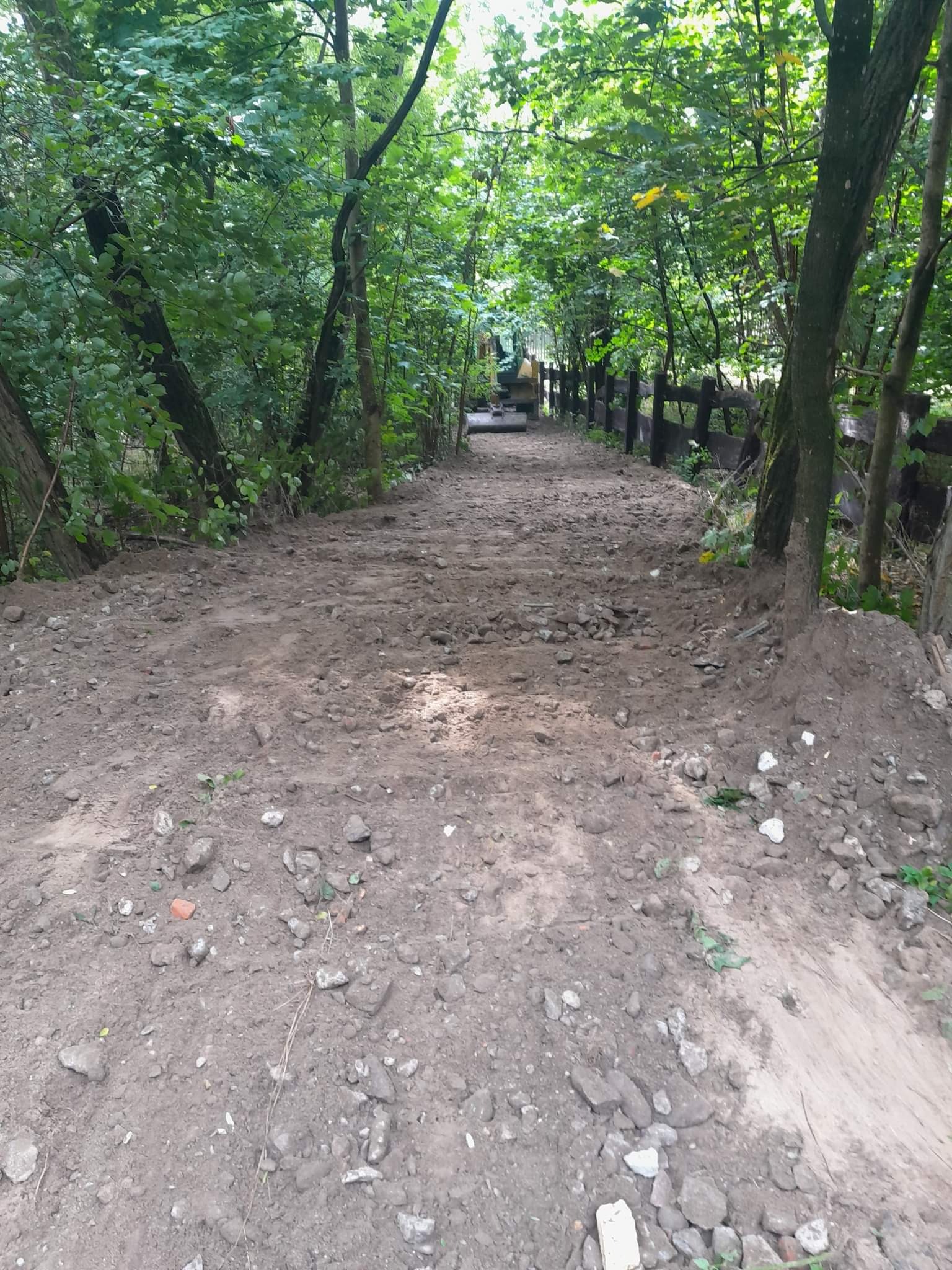 Wyrównany teren pod drogę leśną z widocznym fragmentem ogrodzenia i koparką w oddali.