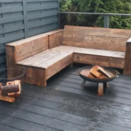 Drewniany narożnik ogrodowy na tarasie z szarej deski kompozytowej, obok kosz z drewnem i palenisko, w tle szare panele ogrodzeniowe i zieleń.