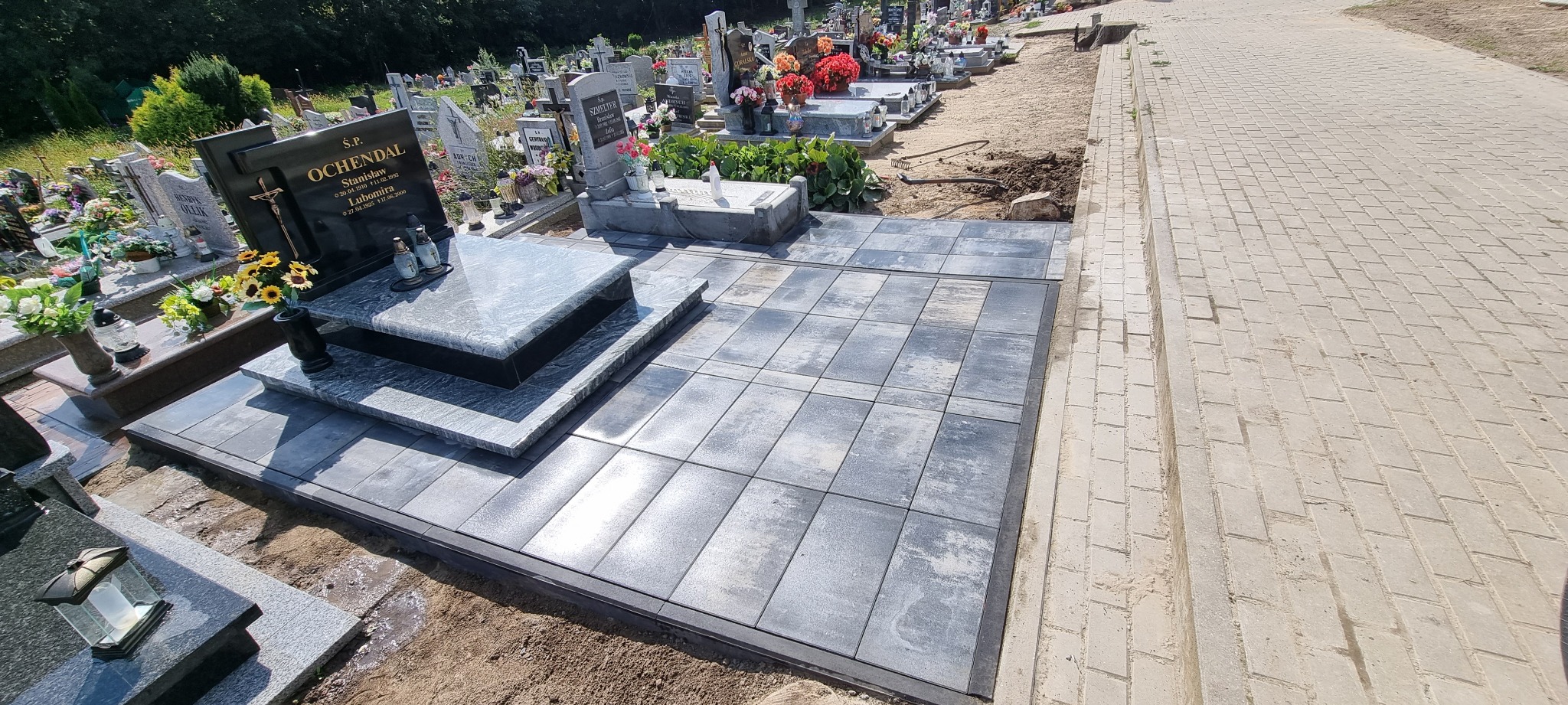 Częściowo ułożona, szara kostka brukowa wokół nagrobka na cmentarzu, widok z góry, jasne oświetlenie.