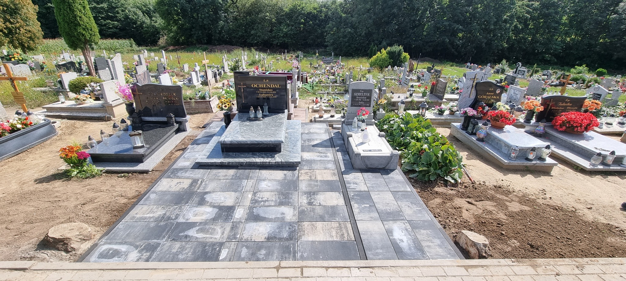 Częściowy widok cmentarza z nowo ułożoną, szarą kostką brukową wokół kilku nagrobków, otoczenie w trakcie prac ziemnych, w tle liczne groby i zieleń.