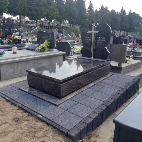 Ciemnoszary nagrobek z granitu z kostką brukową wokół, cmentarz w tle.