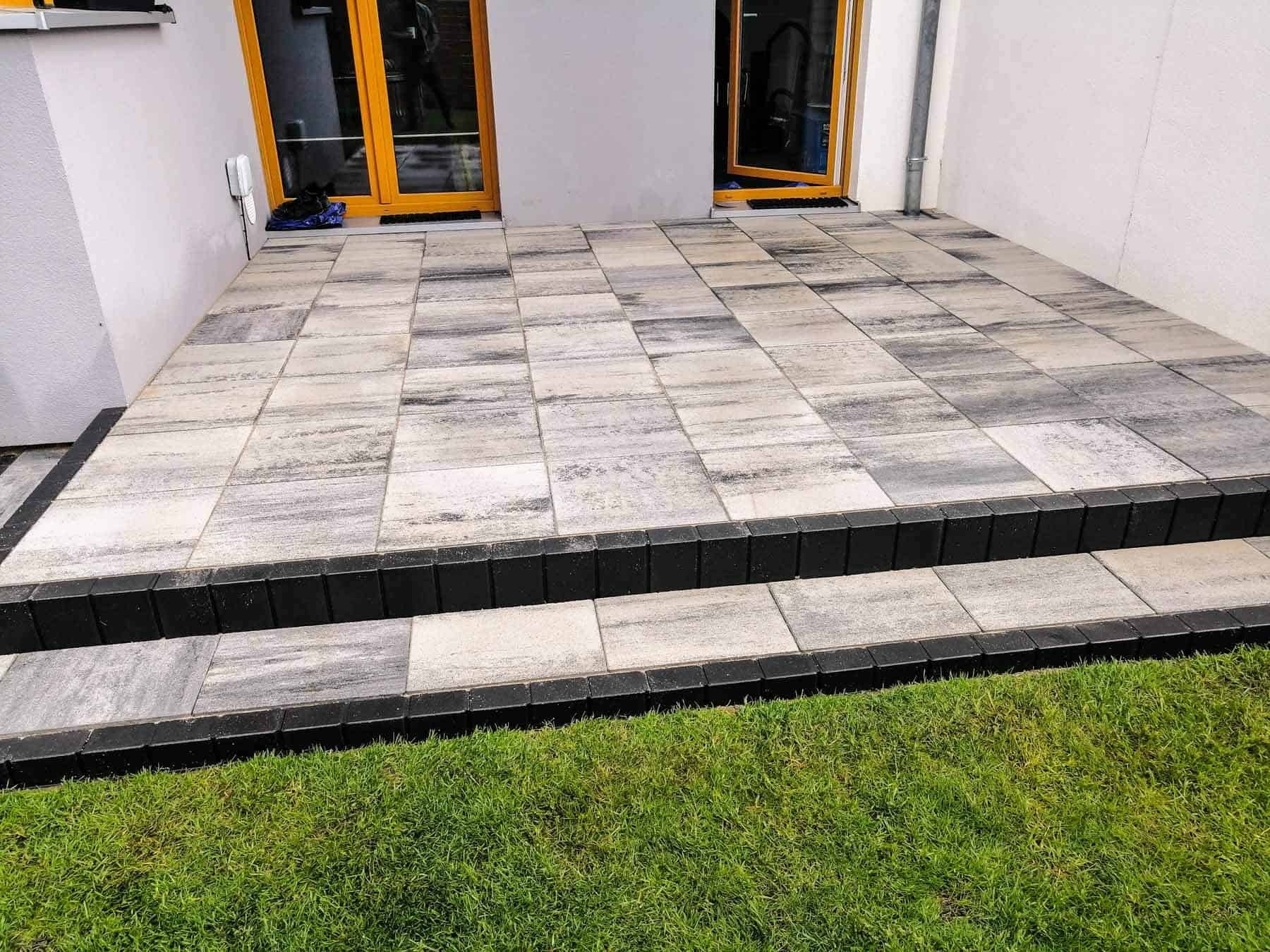 Ułożona kostka brukowa z dwoma stopniami, obrzeżami z ciemnej kostki i trawnikiem w tle, widok z poziomu trawy.