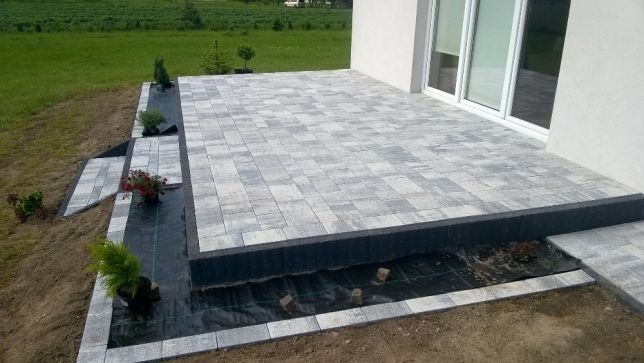 Nowoczesny taras z szarej kostki brukowej ułożonej w prostokątnym wzorze, z czarnym obrzeżem, widok na trawnik i rośliny, z fragmentem białej elewacji budynku.