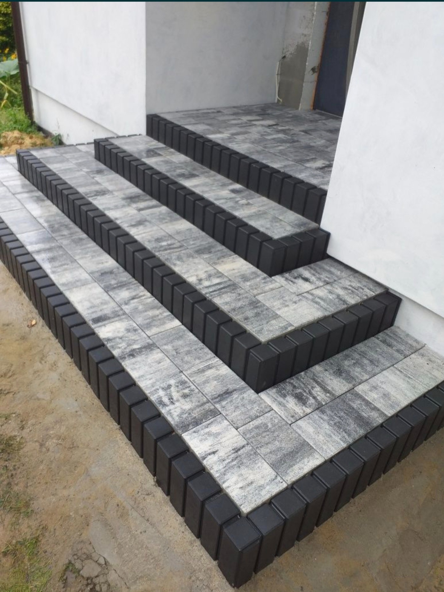 Schody wejściowe wyłożone szarą kostką brukową z czarnym obramowaniem, prowadzące do białego budynku, widoczne niedociągnięcia w wykończeniu terenu.