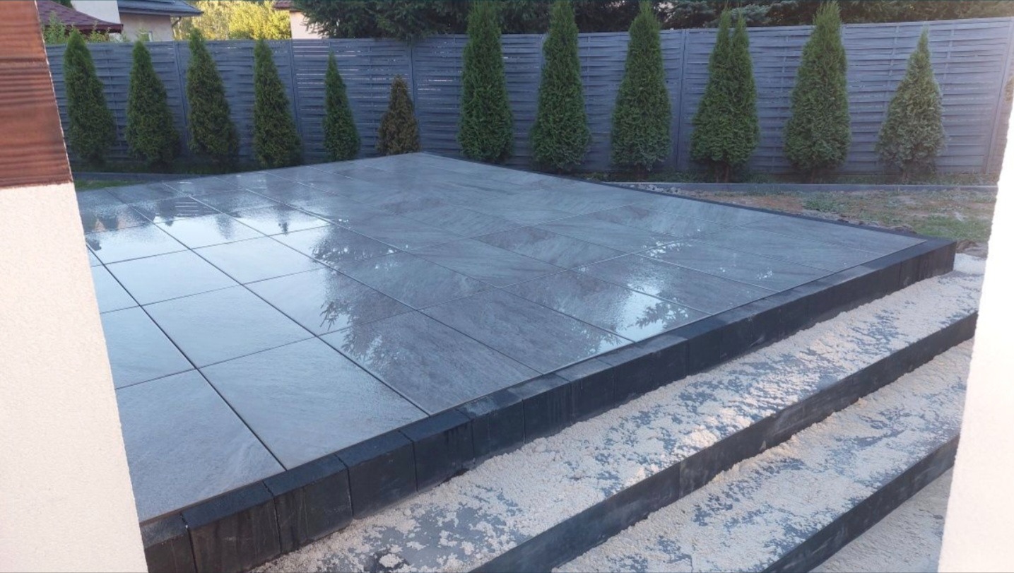 Nowoczesny taras z szarych płyt brukowych, obrzeża z czarnej kostki, trzy stopnie schodów z piaskiem, tło z tujami i drewnianym ogrodzeniem.