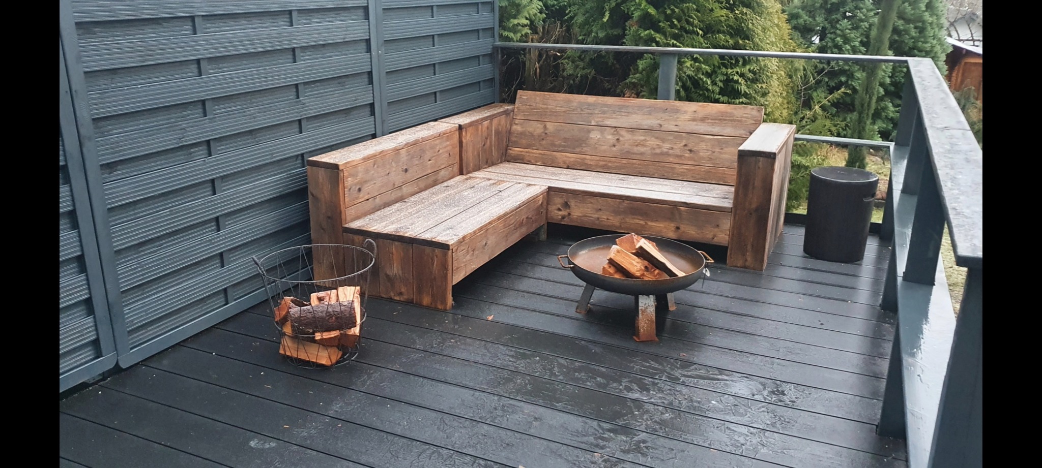 Drewniany narożnik ogrodowy na tarasie z szarej deski kompozytowej, obok kosz z drewnem i palenisko, w tle szare panele ogrodzeniowe i zieleń.