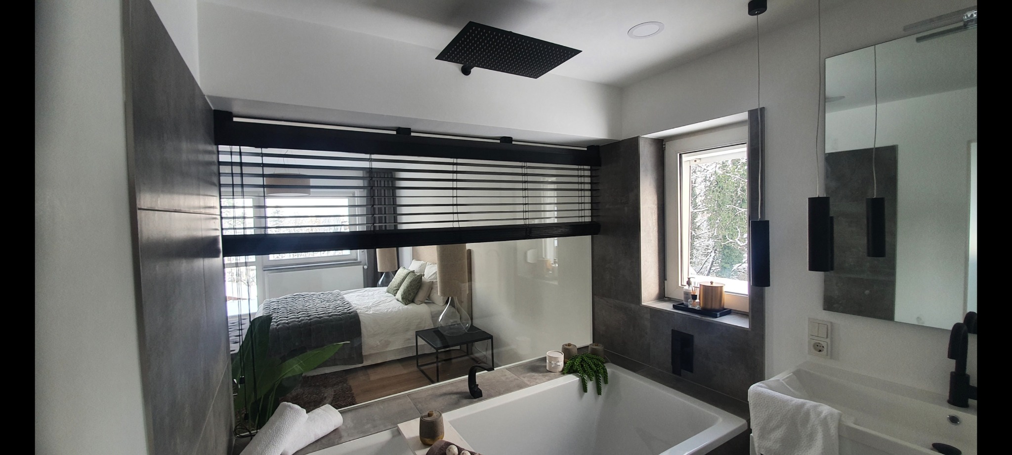 Nowoczesna łazienka z wanną i widokiem na sypialnię przez szklaną ścianę z żaluzjami, czarna armatura, deszczownica, okno z widokiem na zimowy krajobraz.