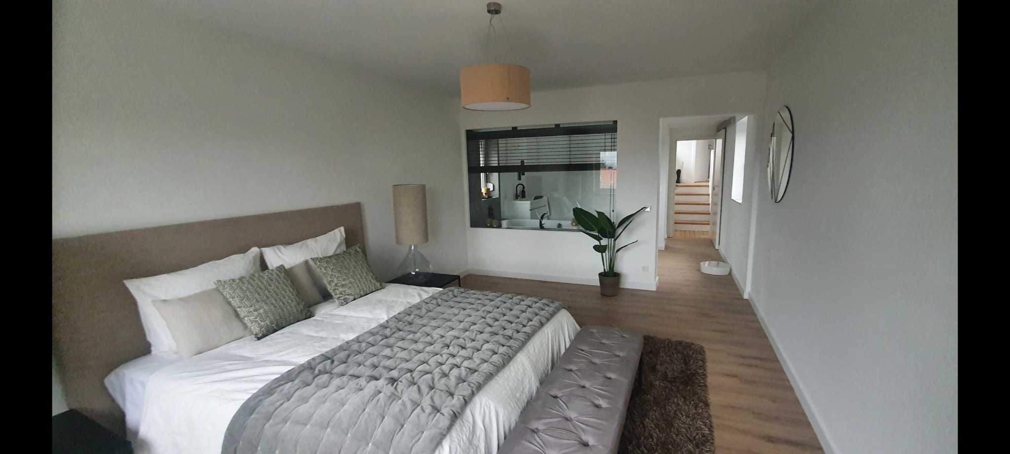 Jasna sypialnia z widokiem na łazienkę przez okno z żaluzjami, drewnianą podłogą i rośliną doniczkową.