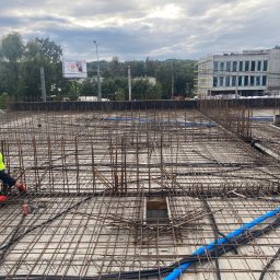 Monolit Paczek - Praca zbrojarza na budowie: zbrojenie stropu budynku żelbetowego z widocznymi elementami instalacji elektrycznej i rurami. W tle budynek w trakcie budowy i reklama.