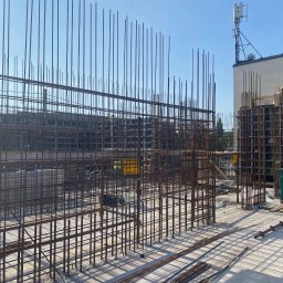 Monolit Paczek - Konstrukcja zbrojeniowa z widocznymi prętami stalowymi na placu budowy, w tle budynek z anteną i pracujący robotnicy.