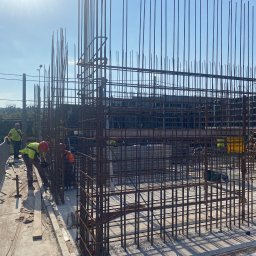 Monolit Paczek - Praca zbrojarzy na budowie: montaż metalowej konstrukcji z prętów zbrojeniowych na tle błękitnego nieba. Widoczni pracownicy w kaskach i kamizelkach.