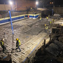 Monolit Paczek - Nocna budowa: Zbrojenie fundamentów pod budynek, pracownicy w kaskach i kamizelkach odblaskowych, oświetlenie budowlane, widok z góry na siatkę zbrojeniową.