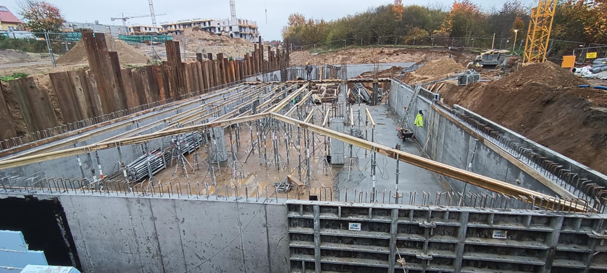 Wykop pod fundamenty z szalunkami i zbrojeniem, widok z góry. Na drugim planie budowa osiedla i dźwig. Metalowe podpory szalunków.