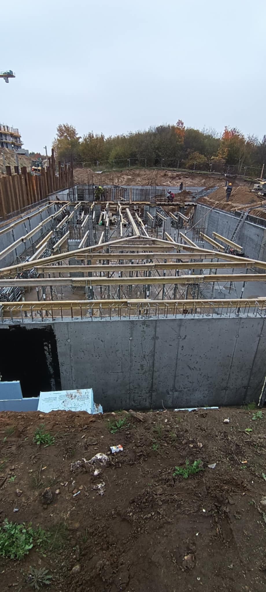 Betonowy fundament z drewnianym szalunkiem i zbrojeniem, widok z góry na placu budowy z zielenią w tle, pochmurne niebo.