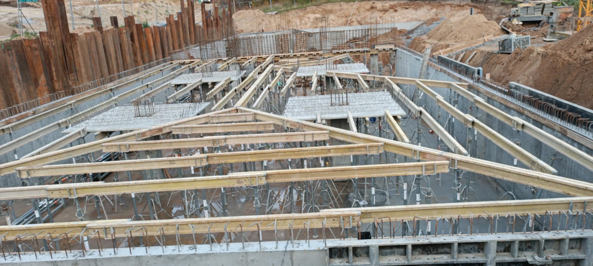 Widok z góry na fundamenty budynku w trakcie budowy, z drewnianymi belkami i stalowymi elementami zbrojenia. W tle widoczne ziemne skarpy i metalowe ścianki.