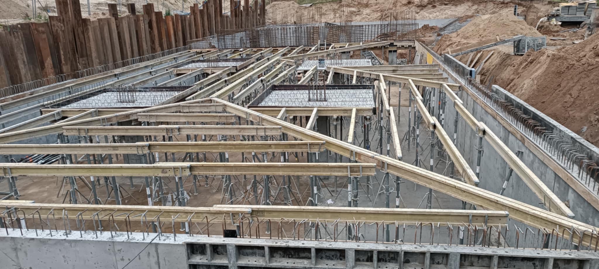Skomplikowane zbrojenie i szalunki fundamentów pod budynek z widocznymi elementami stalowymi i drewnianymi, przygotowane do zalania betonem.