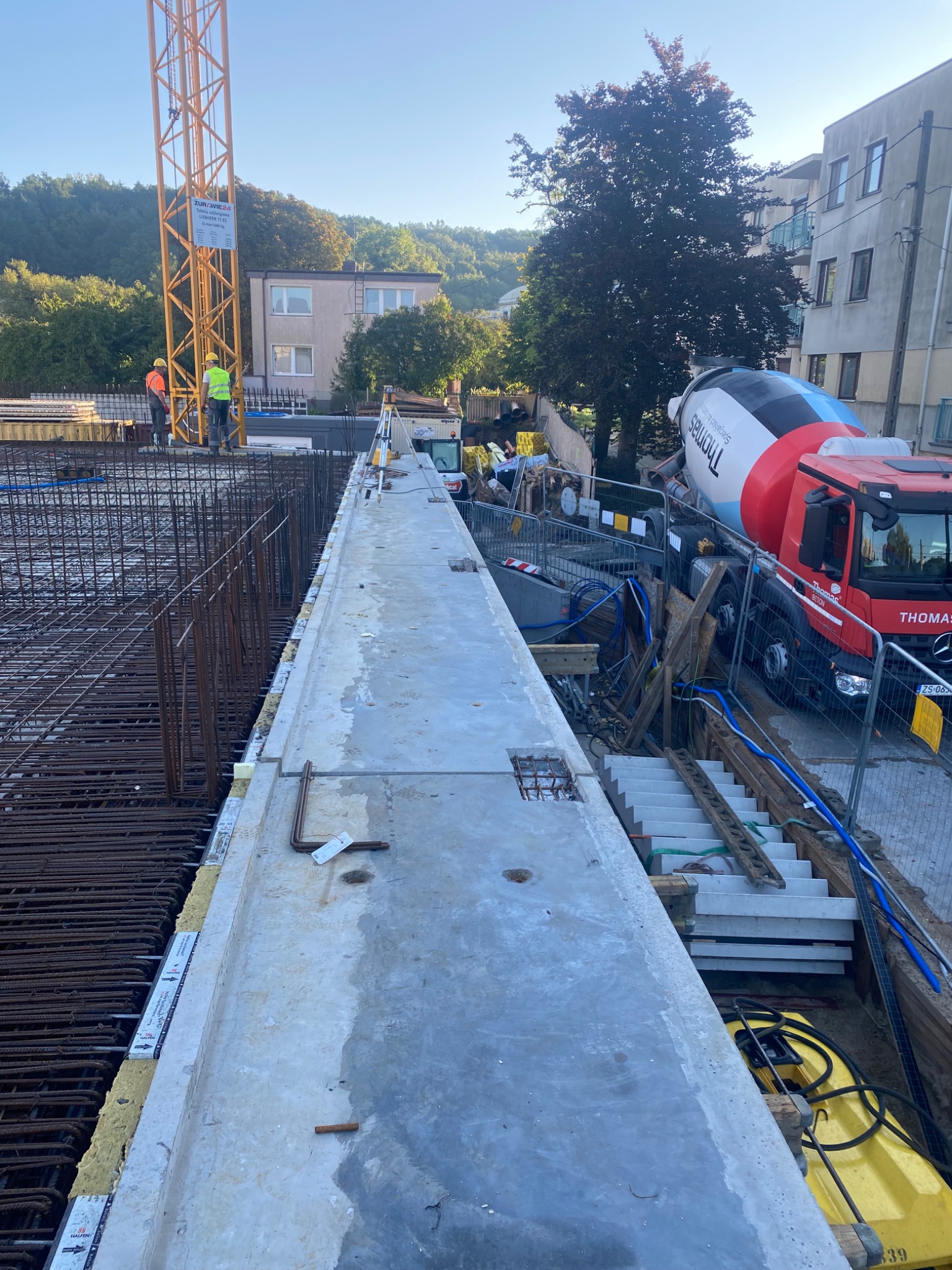 Betoniarka Thomas i zbrojenie na budowie budynku żelbetowego. Widoczny dźwig i pracownicy w odblaskowych kamizelkach. W oddali budynki i drzewa.