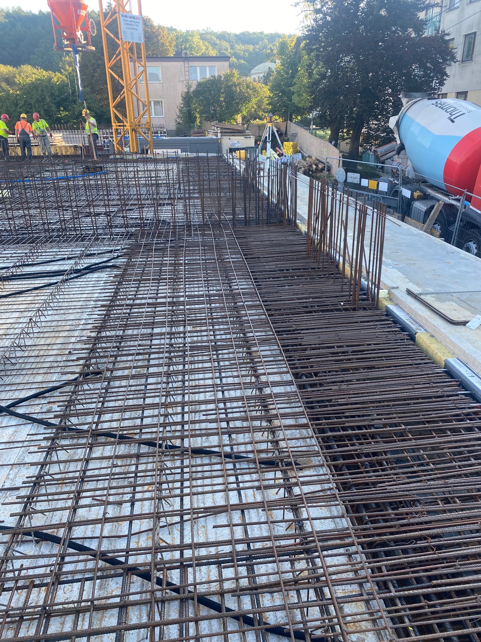 Siatka zbrojeniowa na budowie, widok z góry. Żuraw budowlany w tle, betoniarka w prawym górnym rogu. Prace zbrojarskie w toku, przygotowanie do betonowania stropu.