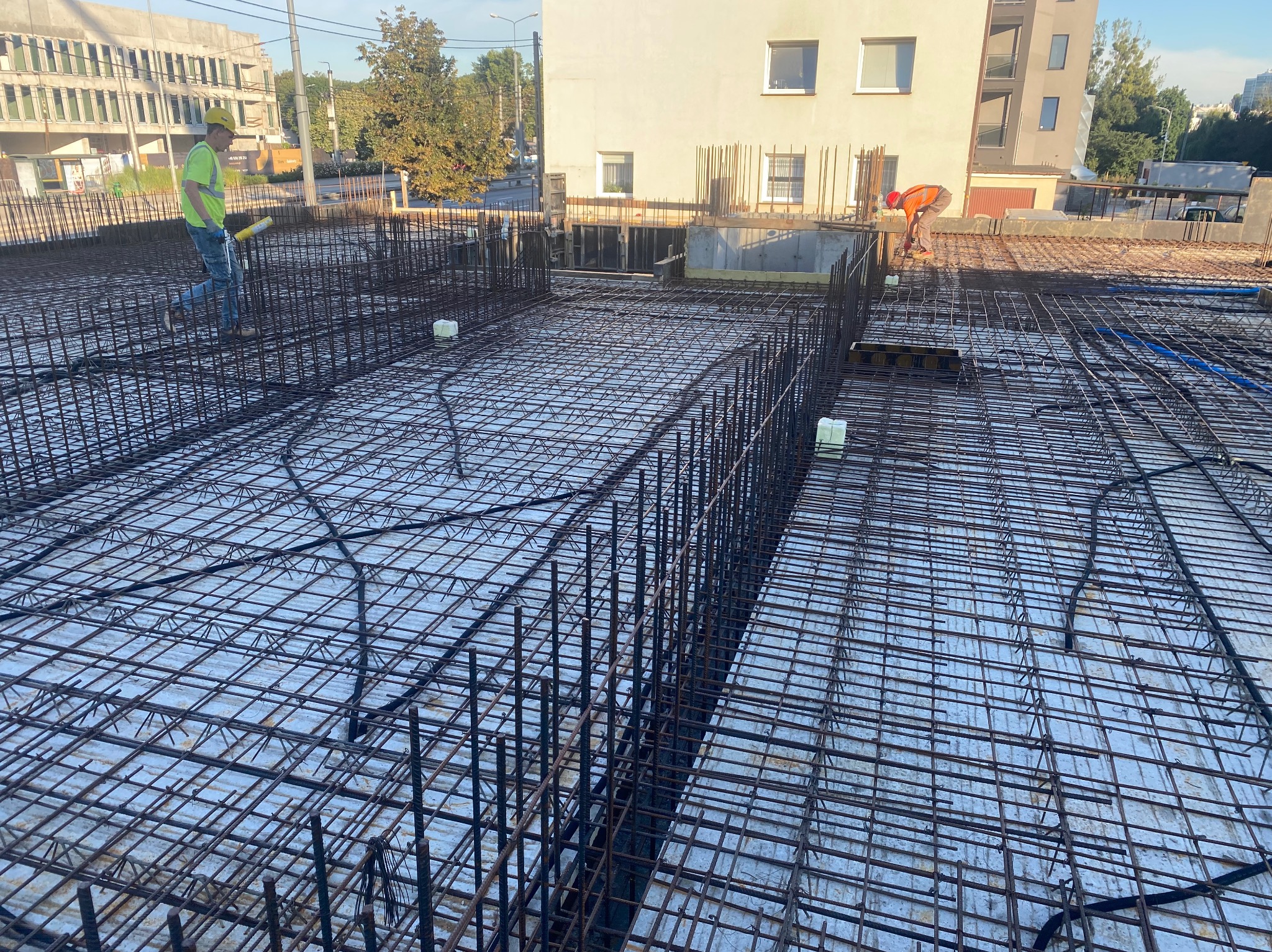 Siatka zbrojeniowa na budowie z pracownikami w kaskach i odblaskowych kamizelkach, przygotowana do zalania betonem. Widok z góry, w tle budynki.