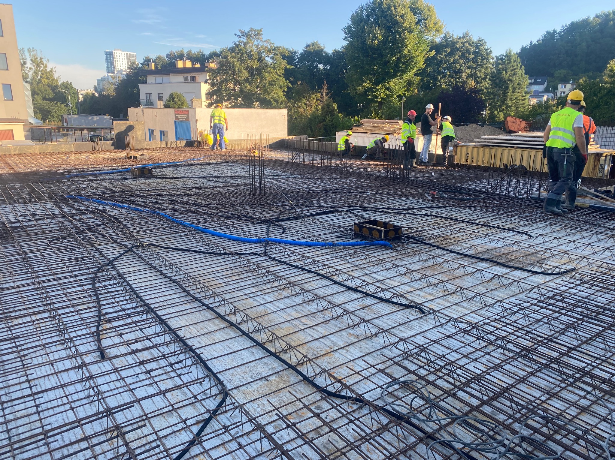 Rozległe zbrojenie stropu z widocznymi niebieskimi rurami instalacji, ekipa budowlana w tle, słoneczny dzień.