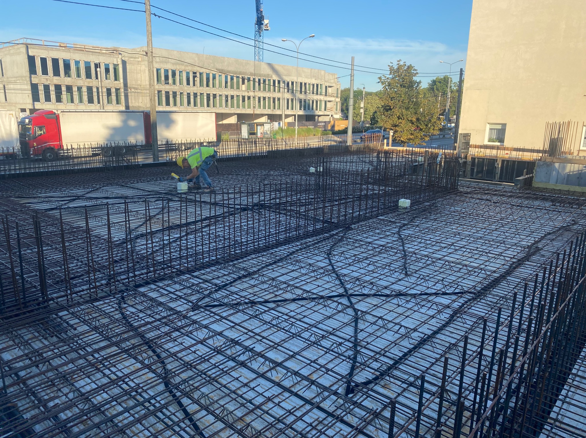Zbrojenie płyty fundamentowej budynku w trakcie prac. Widoczny pracownik w żółtej kamizelce i ułożone pręty zbrojeniowe. W tle budynek w stanie surowym otwartym.