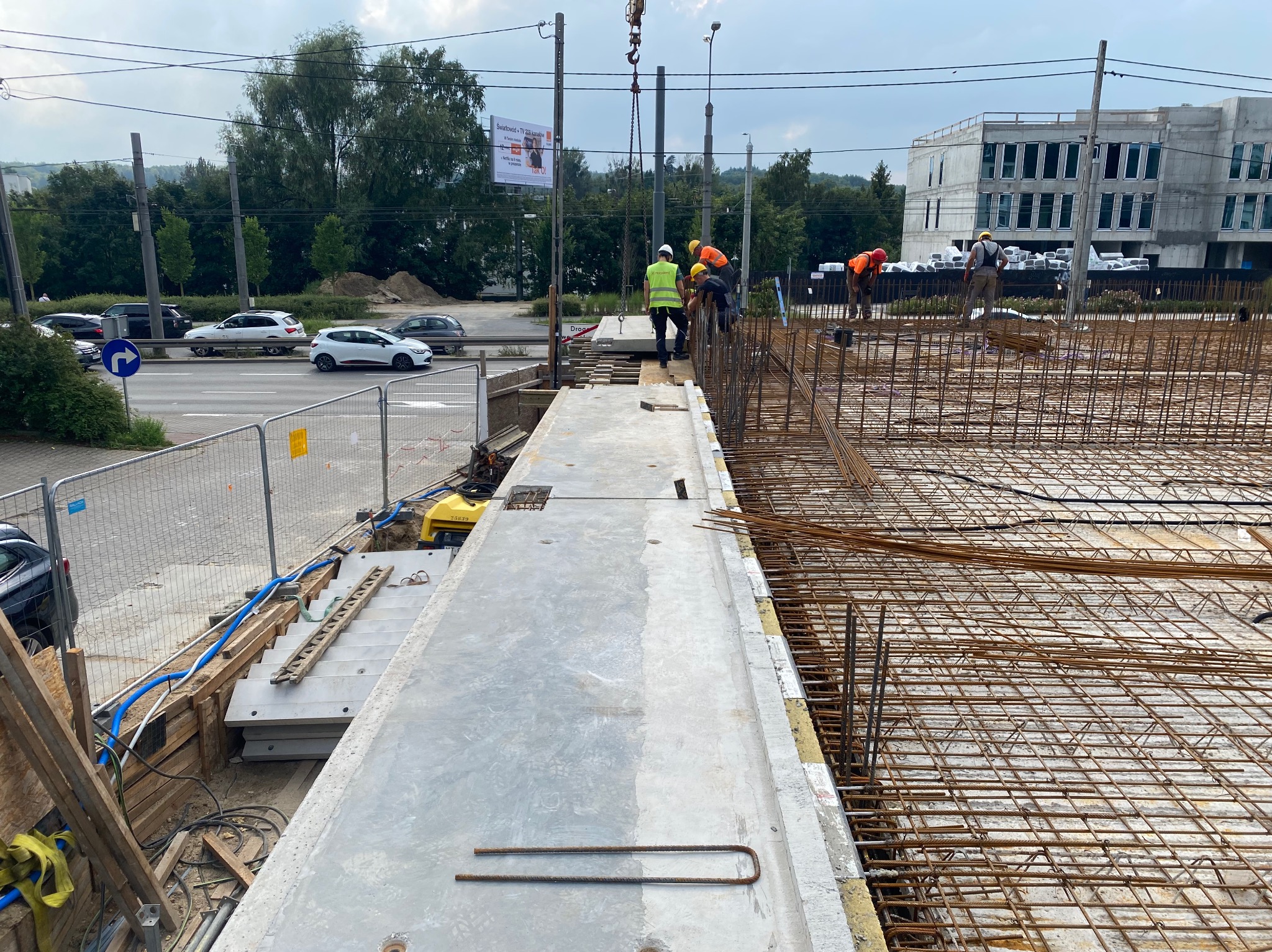 Budowa budynku żelbetowego: montaż prefabrykowanych płyt stropowych dźwigiem. Widoczna konstrukcja zbrojeniowa, pracownicy w kaskach i kamizelkach. W tle budynek w stanie surowym.