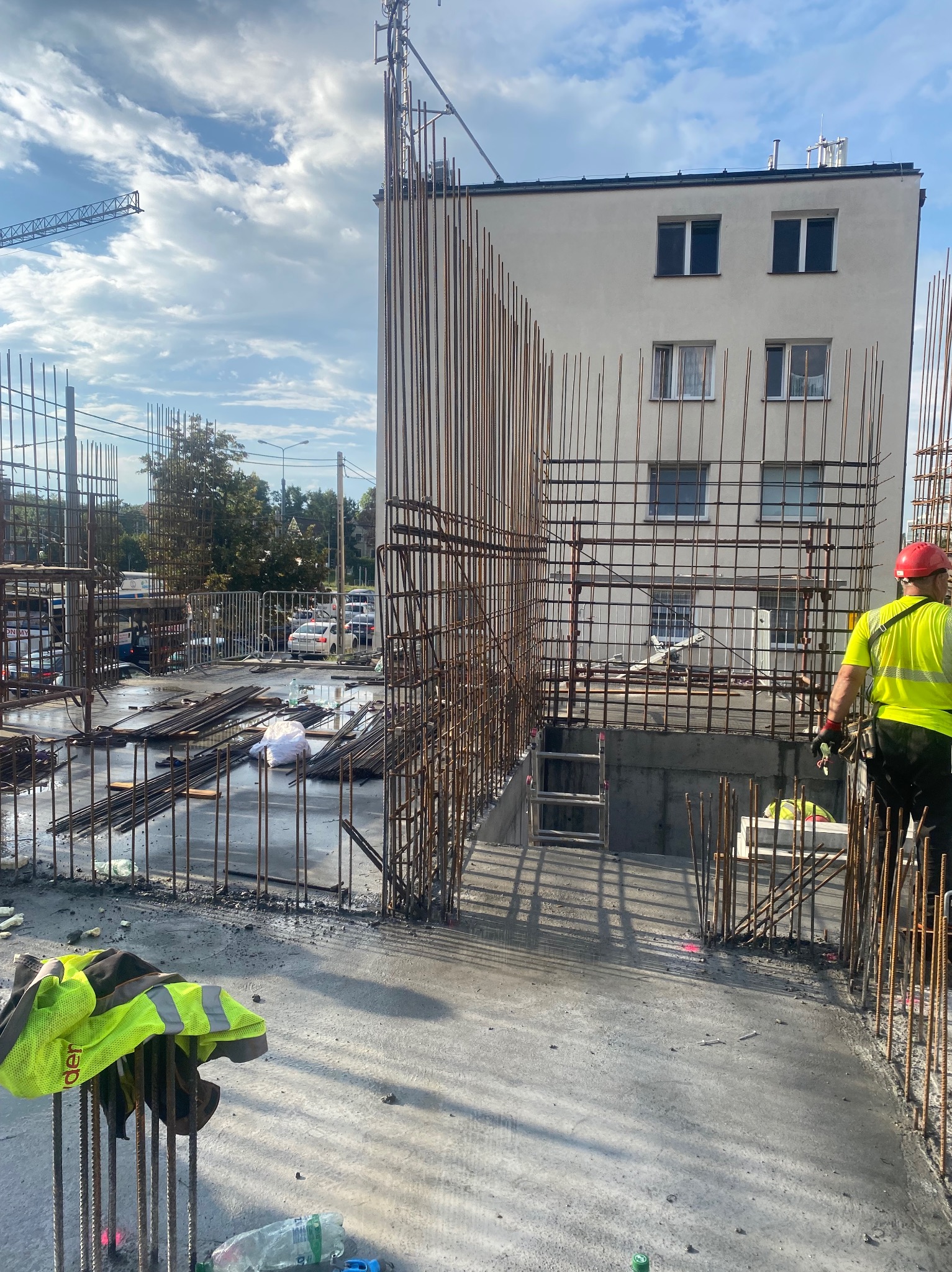 Zbrojenie pionowe i poziome na budowie, w tle budynek z białej cegły. Widoczny pracownik w odblaskowej kamizelce i czerwonym kasku.