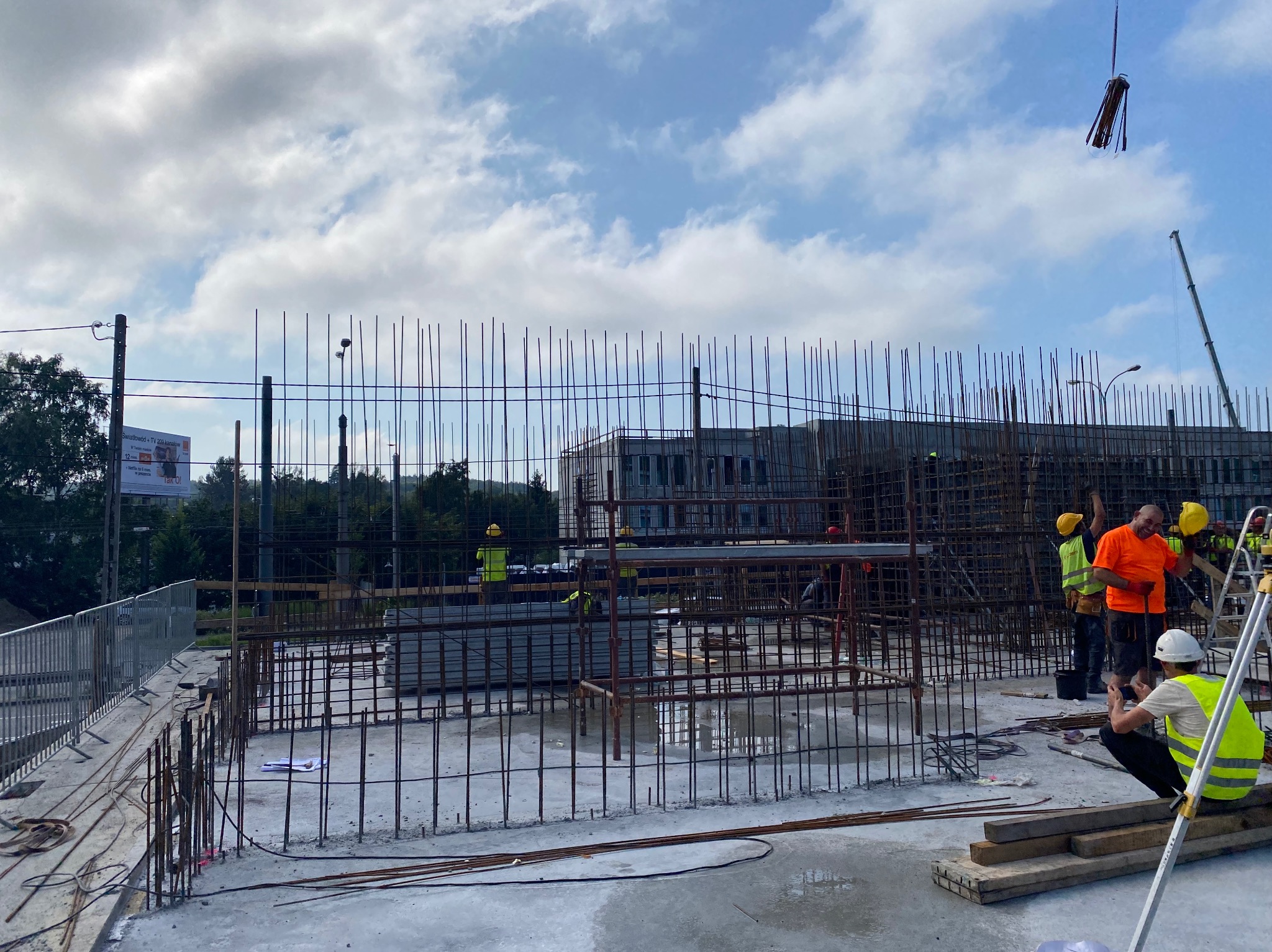 Zbrojenie płyty fundamentowej budynku w trakcie budowy. Widoczni pracownicy w kaskach i kamizelkach odblaskowych. Żuraw podnosi elementy konstrukcyjne.