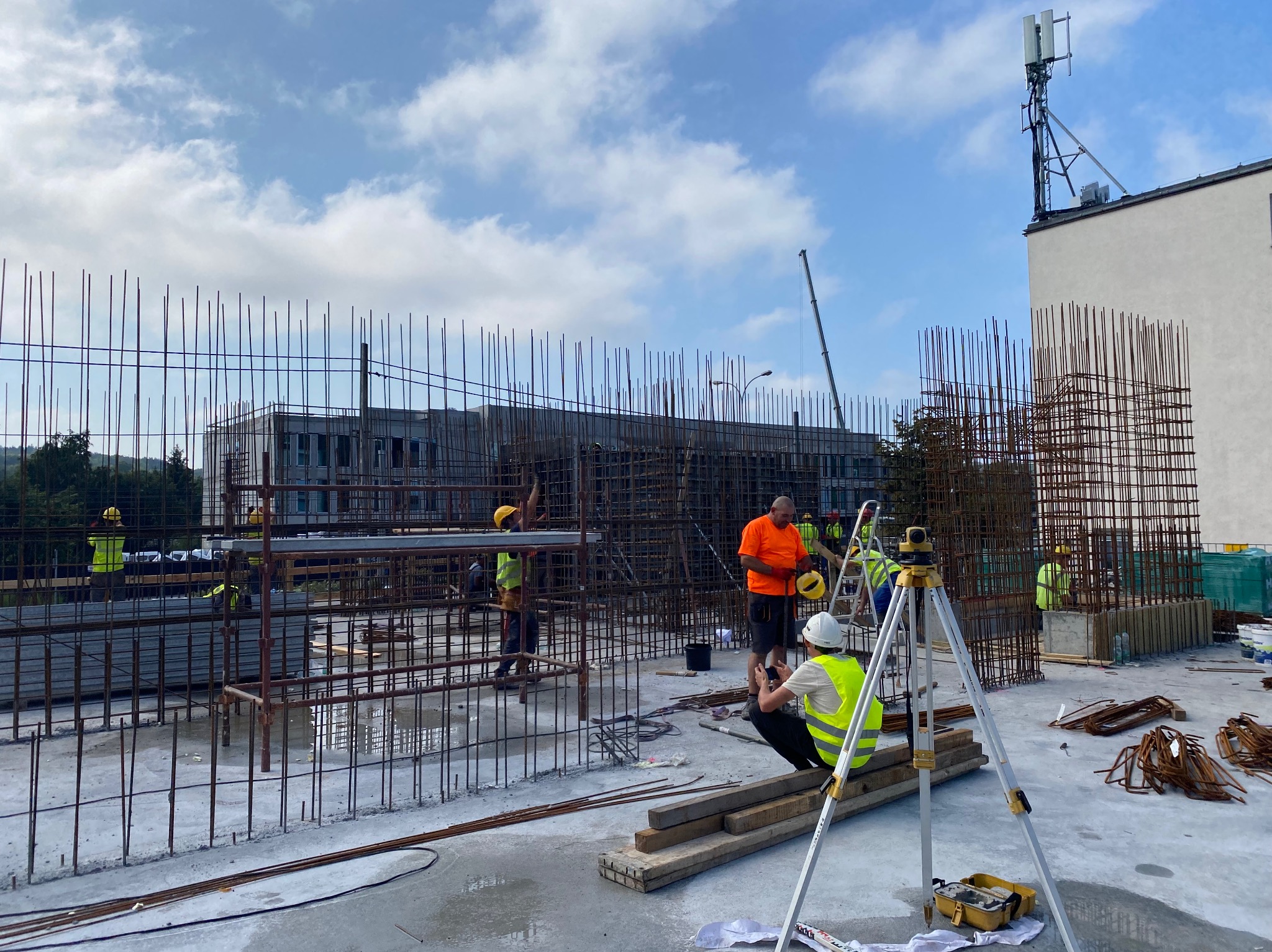 Ekipa budowlana w trakcie prac zbrojarskich na budowie budynku żelbetowego. Widoczne zbrojenia, pracownicy w kaskach i kamizelkach oraz instrument geodezyjny.