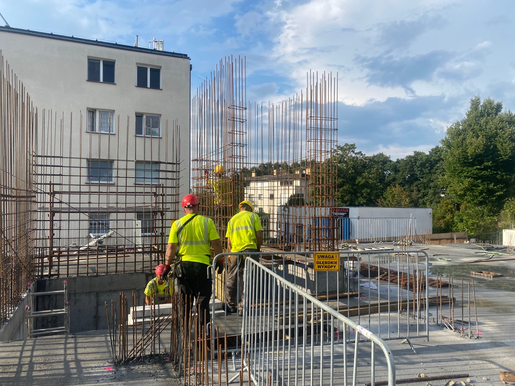 Prace zbrojarskie na budowie z widoczną konstrukcją z prętów stalowych i pracownikami w odblaskowych ubraniach. W tle budynek mieszkalny i błękitne niebo.