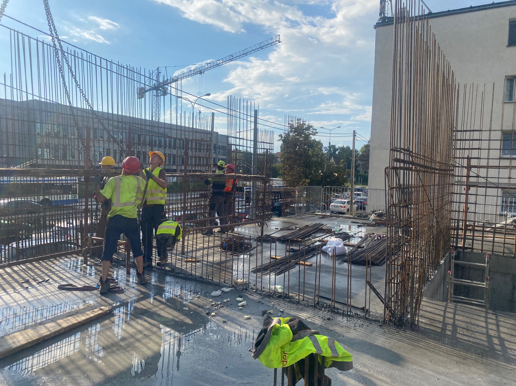Ekipa budowlana w żółtych kamizelkach montuje zbrojenie na placu budowy; w tle budynek i żuraw. Elementy metalowe odbijają światło słoneczne.