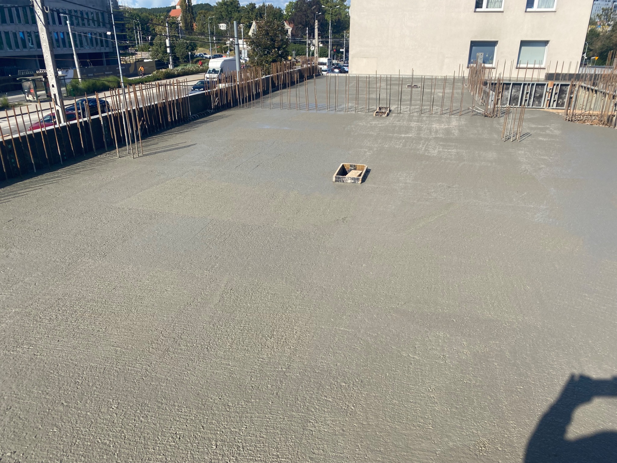 Wylany beton na stropie z wystającymi prętami zbrojeniowymi w tle zabudowa miejska i drzewa. Widoczny fragment cienia osoby robiącej zdjęcie.