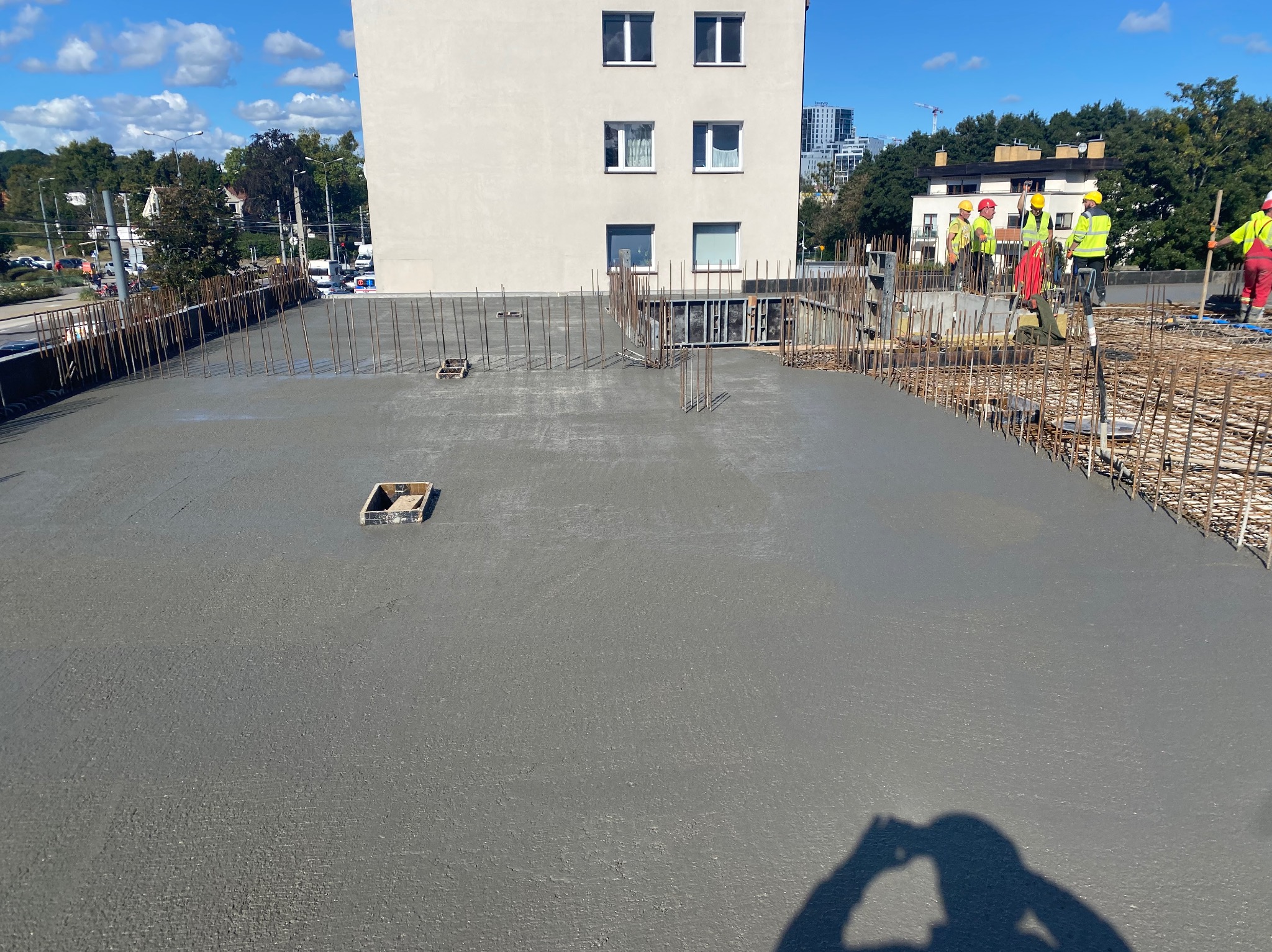 Świeżo wylany beton na budowie z widocznym zbrojeniem i pracownikami w oddali, budynek w tle, słoneczny dzień. Shadow osoby robiącej zdjęcie w rogu kadru.