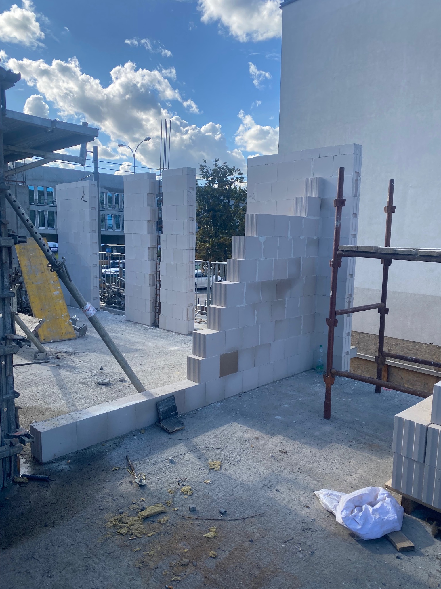 Wznoszenie ścian z białych bloczków na budowie. Widoczne rusztowania, niebo z chmurami w tle. Częściowo wymurowana ściana z widocznymi otworami okiennymi.