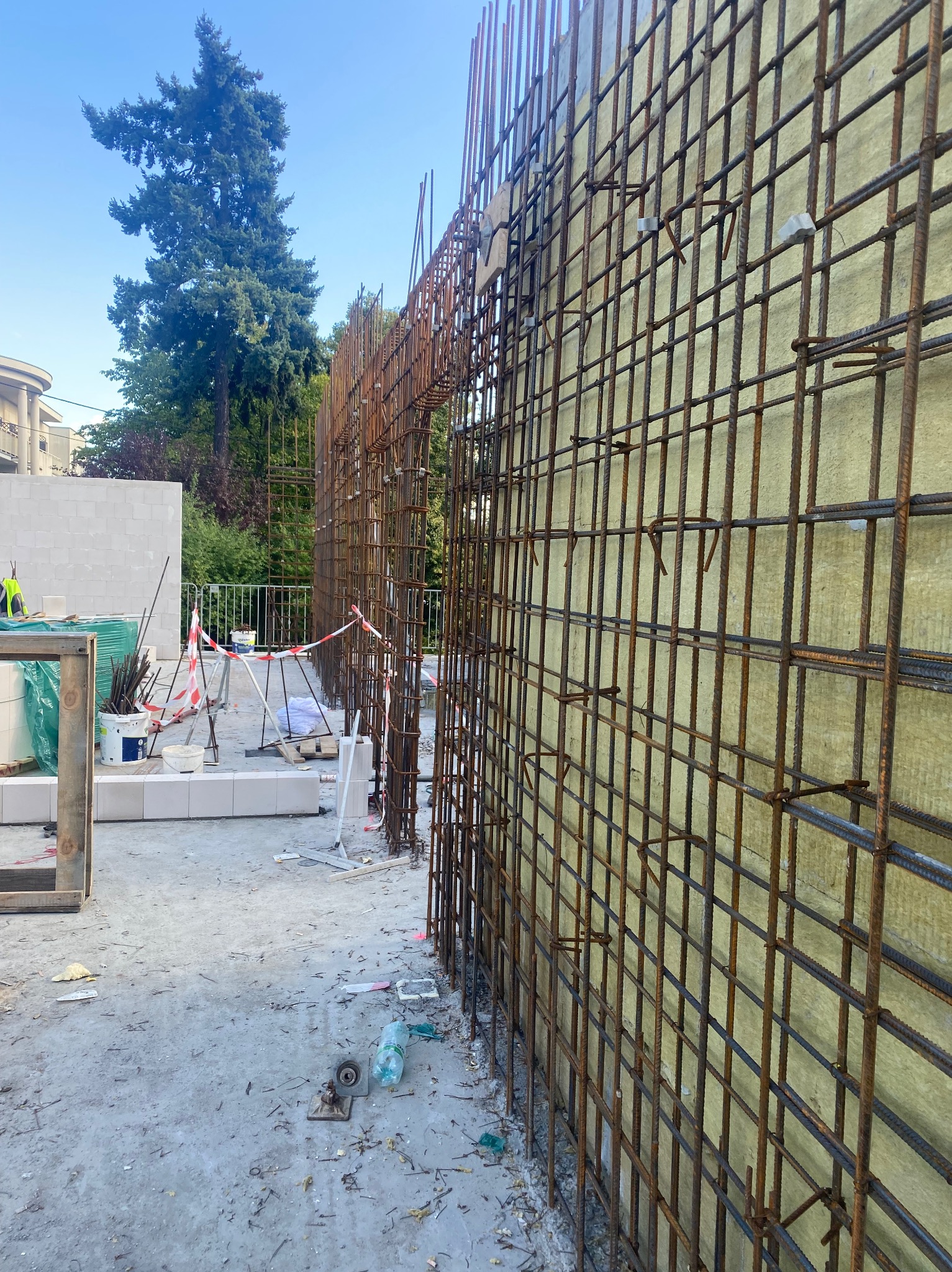 Zbrojenie ściany z żółtą izolacją na budowie. Widoczne stalowe pręty, elementy mocujące i narzędzia. W tle drzewa i budynek.