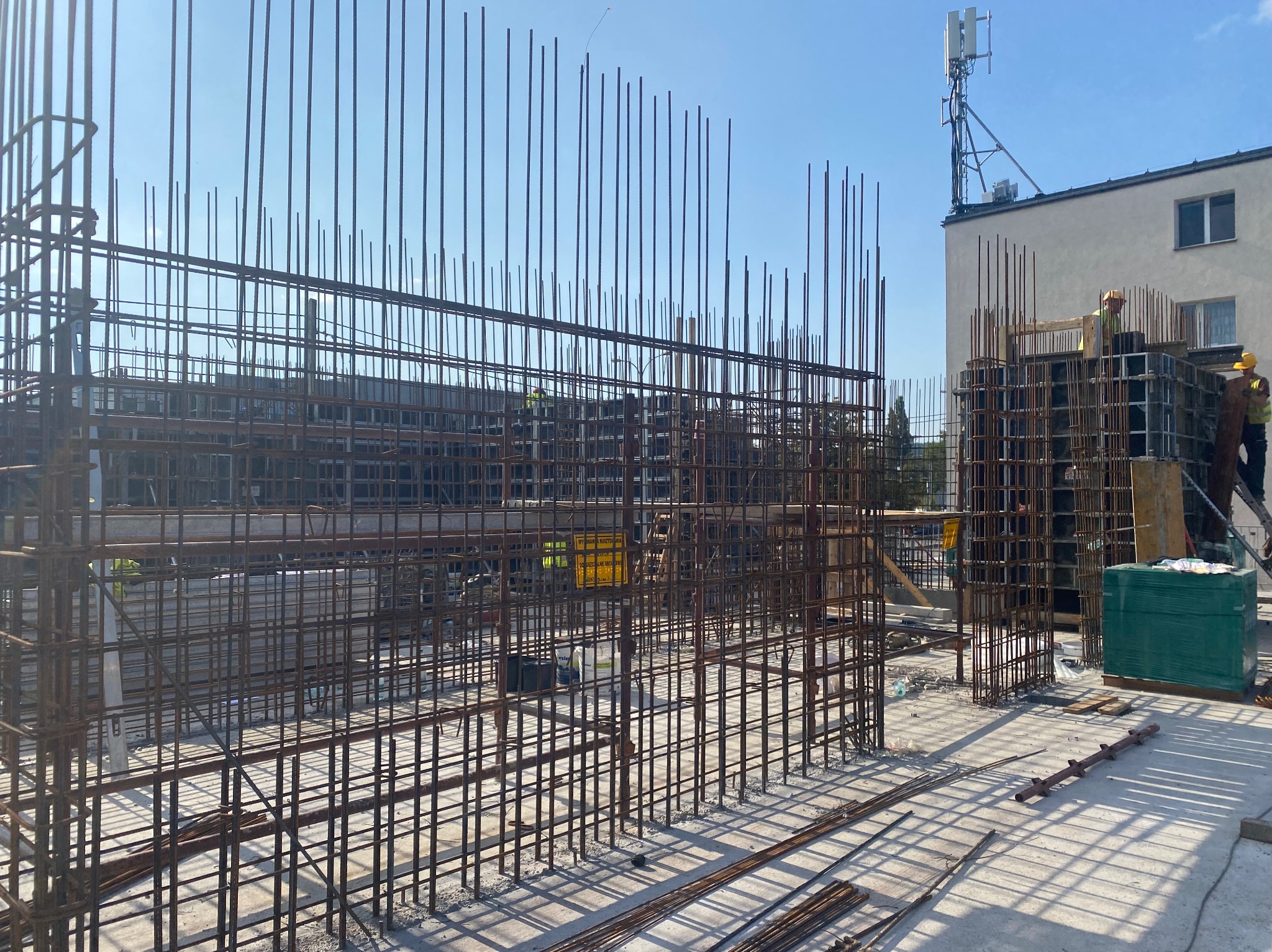 Konstrukcja zbrojeniowa z widocznymi prętami stalowymi na placu budowy, w tle budynek z anteną i pracujący robotnicy.