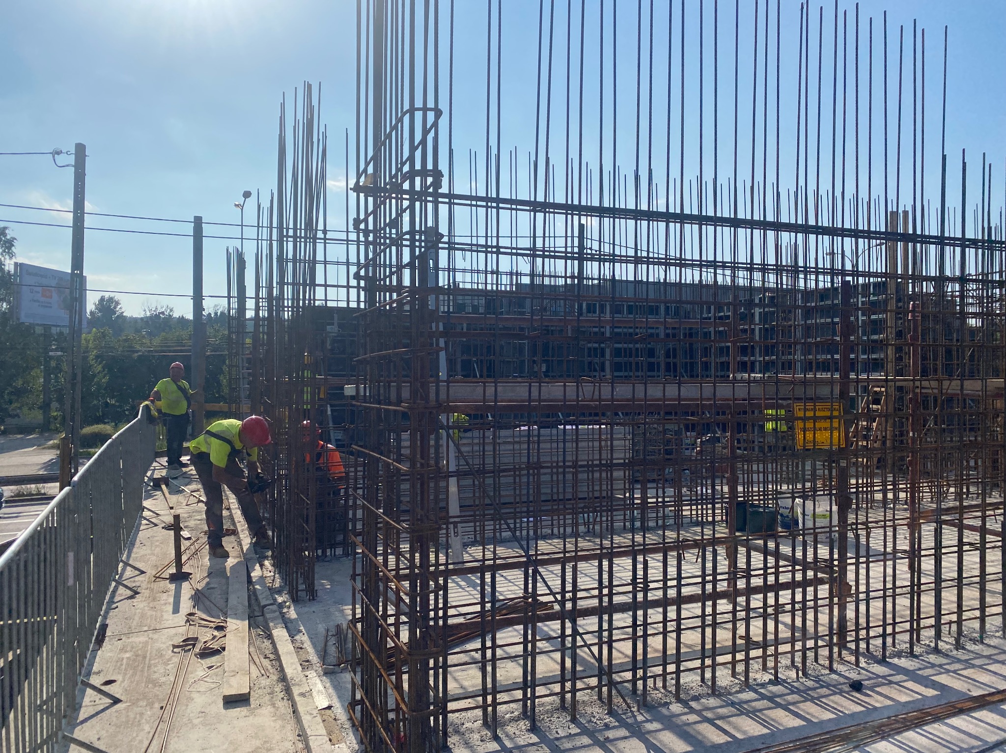 Praca zbrojarzy na budowie: montaż metalowej konstrukcji z prętów zbrojeniowych na tle błękitnego nieba. Widoczni pracownicy w kaskach i kamizelkach.