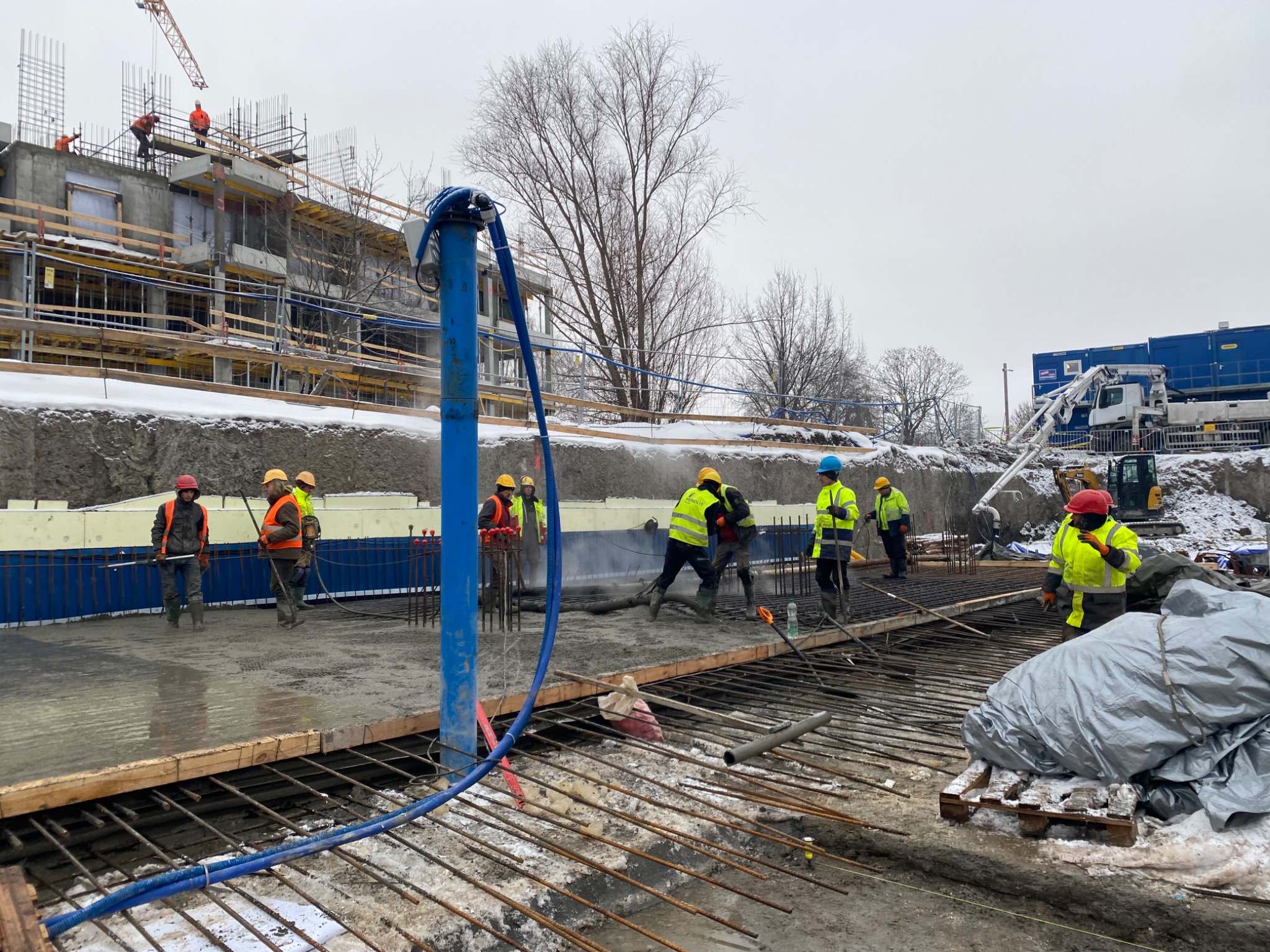 Wylewka betonu na zbrojeniu fundamentów budynku wielorodzinnego. Pracownicy w żółtych kamizelkach i kaskach. W tle budynek w trakcie budowy i dźwig.