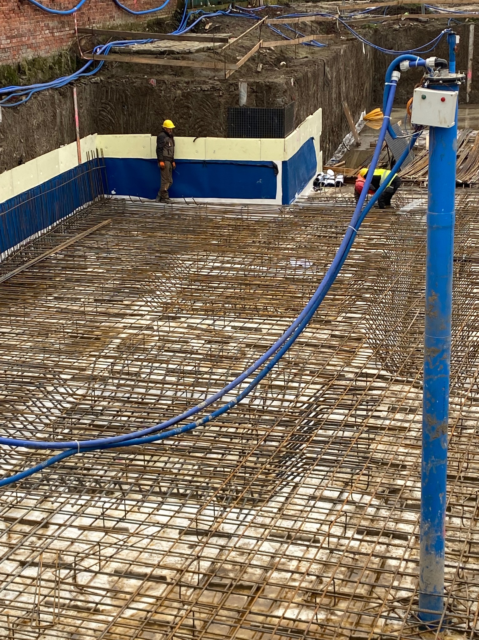 Siatka zbrojeniowa fundamentu z hydroizolacją, widoczny pracownik w żółtym kasku i niebieskie węże. Ujęcie z góry na plac budowy.