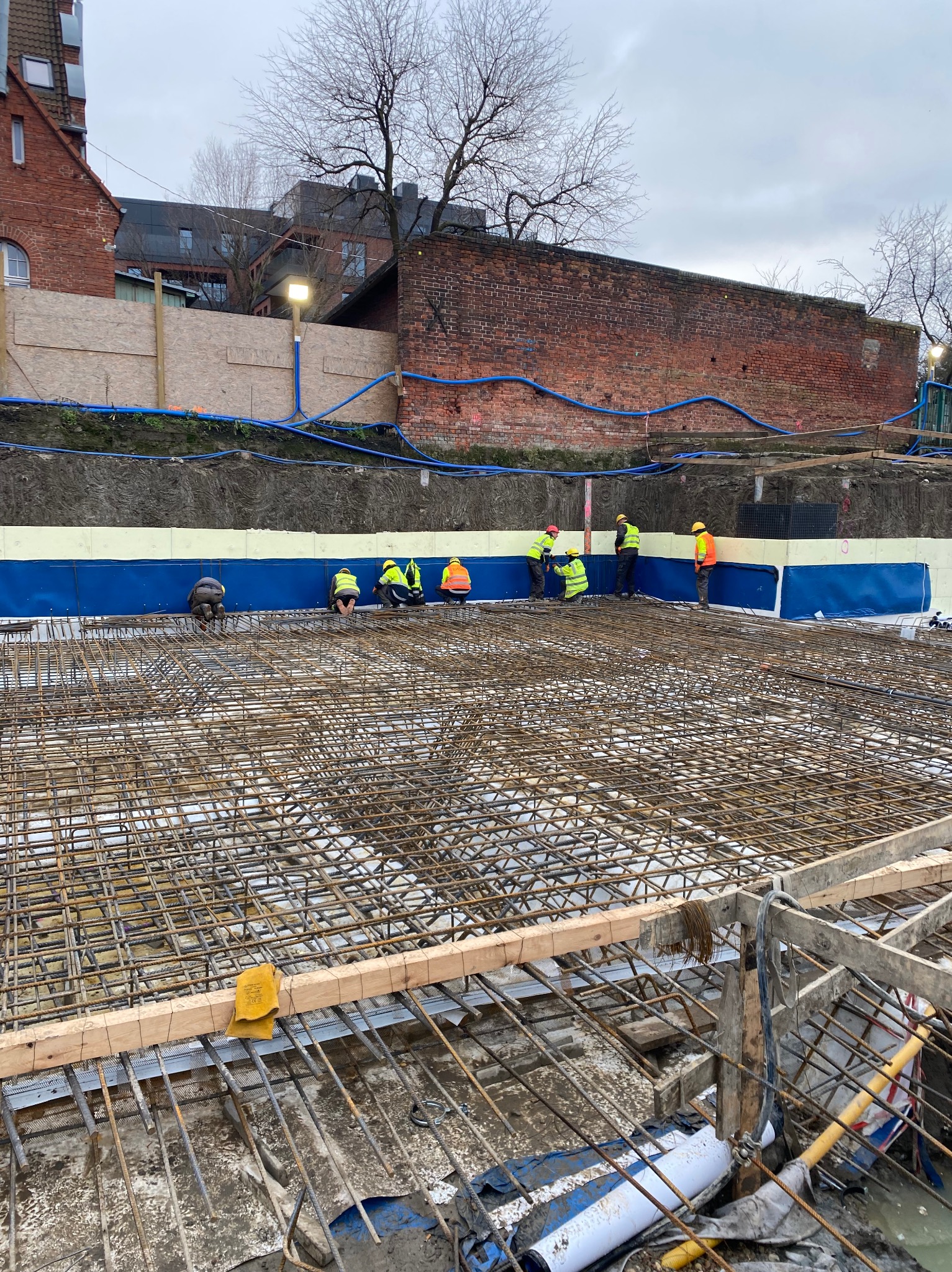 Zbrojenie fundamentów pod budynek z widocznymi pracownikami w odblaskowych kamizelkach. Zbrojenie wykonane na tle ceglanego budynku i nieba.