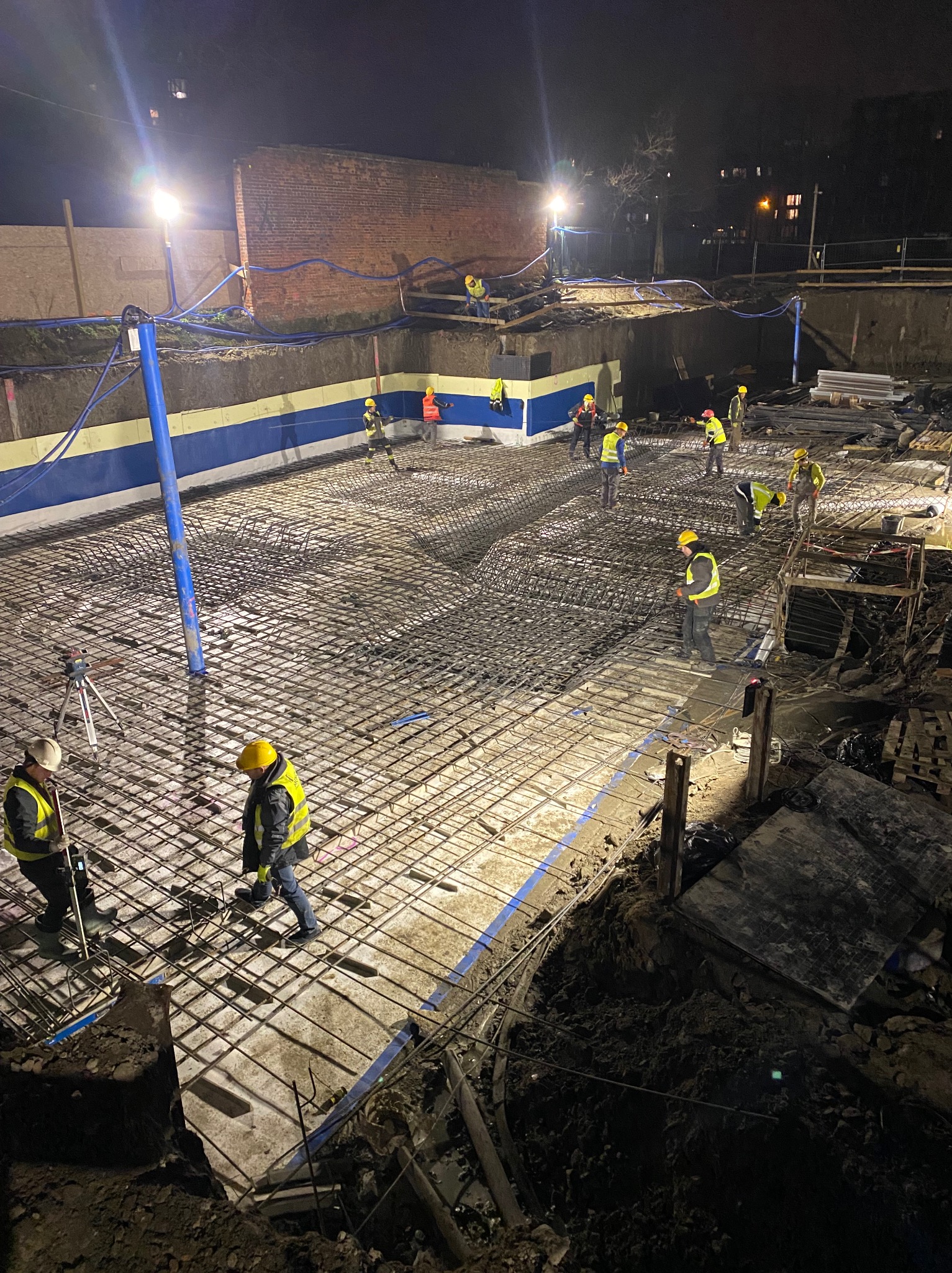 Nocna budowa: Zbrojenie fundamentów pod budynek, pracownicy w kaskach i kamizelkach odblaskowych, oświetlenie budowlane, widok z góry na siatkę zbrojeniową.