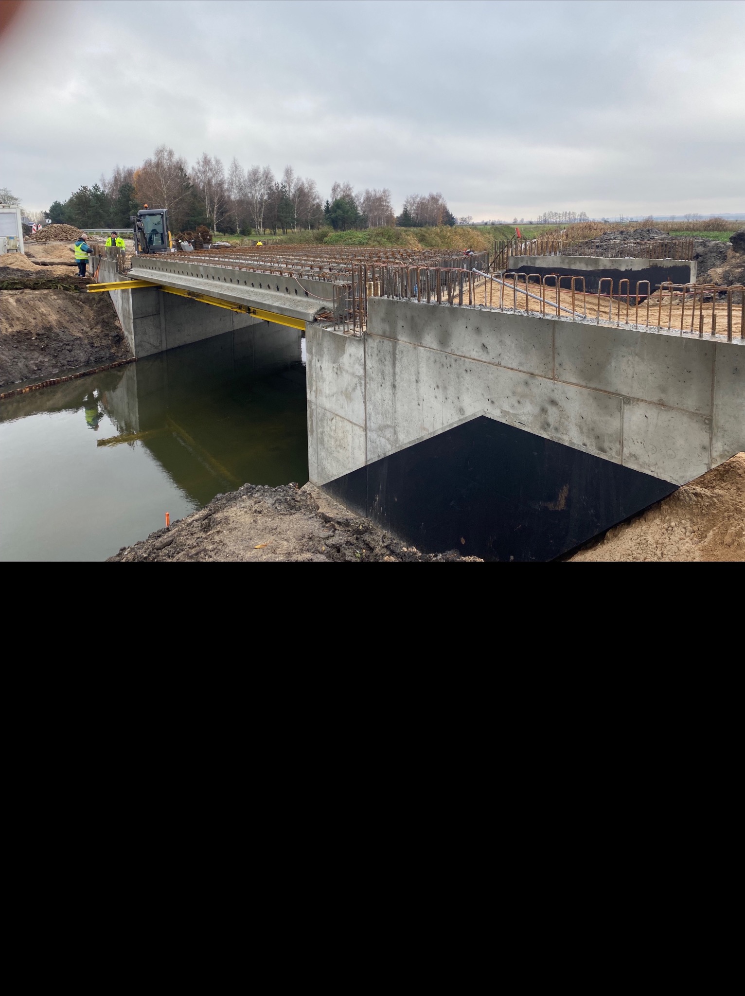 Betonowy most w trakcie budowy nad rzeką, widoczne zbrojenia i pracownicy w odblaskowych kamizelkach. Pochmurne niebo w tle.
