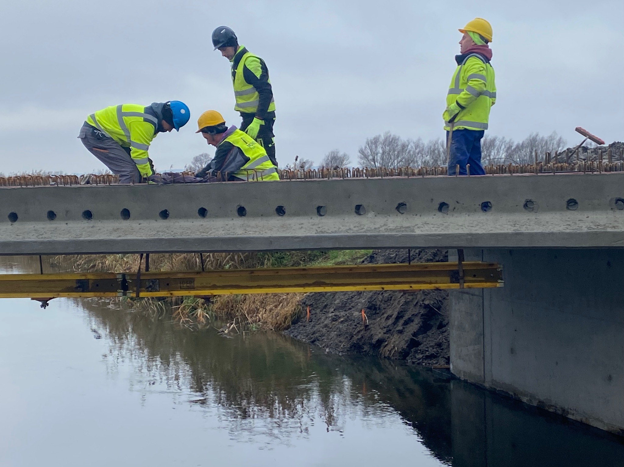Betonowy most w budowie nad rzeką. Trzech robotników w kaskach i kamizelkach pracuje nad zbrojeniem. Widoczne podpory i odbicie w wodzie. Pochmurne niebo.