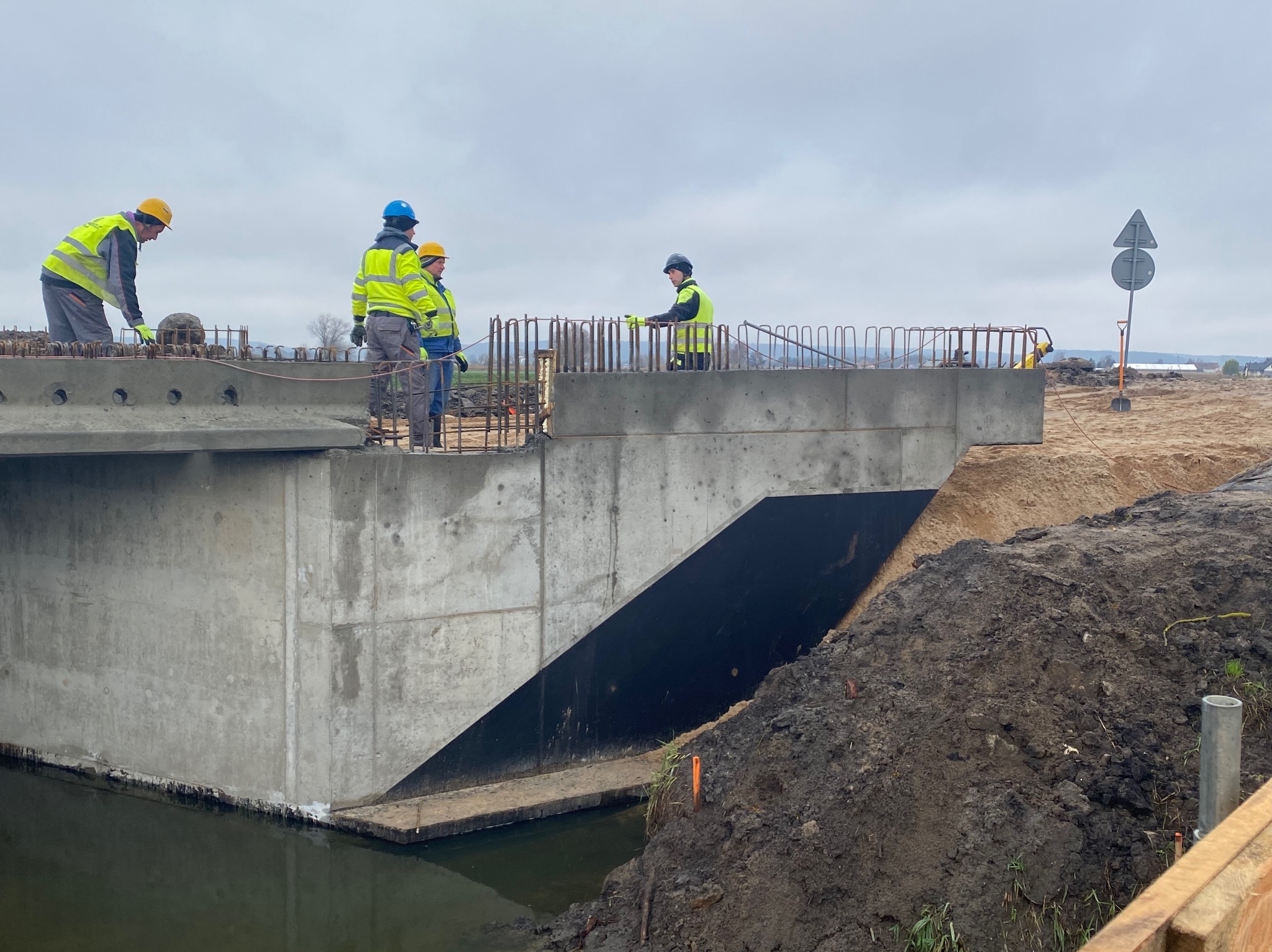 Betonowy most w trakcie budowy nad ciekiem wodnym. Widoczni robotnicy w kaskach i kamizelkach odblaskowych. Zbrojenie na górze konstrukcji. Pochmurne niebo.