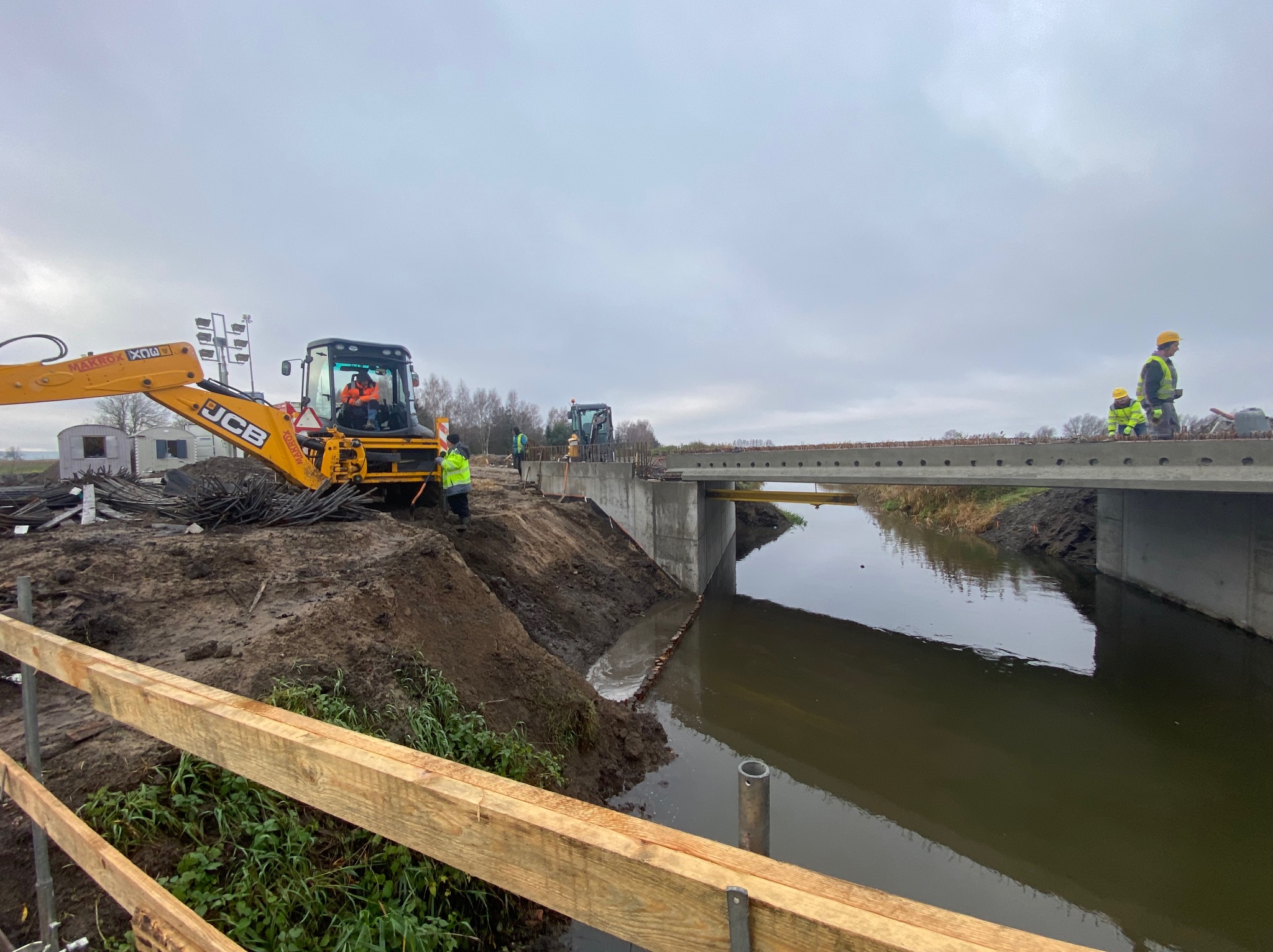 Budowa betonowego mostu nad rzeką. Na pierwszym planie żółta koparka JCB i pracownicy w kaskach. W tle widoczny drugi most i maszyny budowlane.