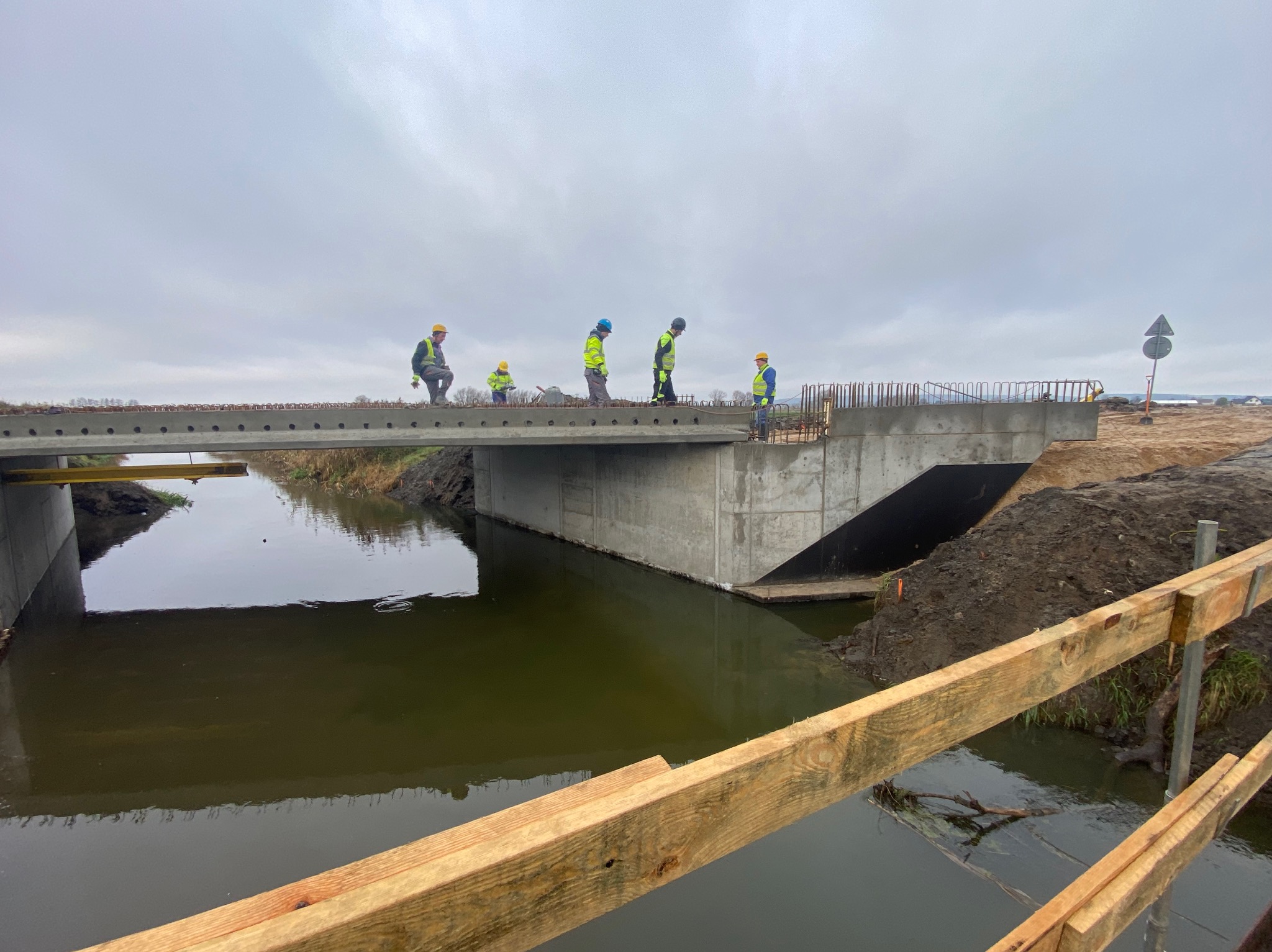 Budowa betonowego mostu nad rzeką. Widoczni pracownicy w kaskach i kamizelkach odblaskowych. Zbrojenie na górze konstrukcji. Pochmurne niebo w tle.